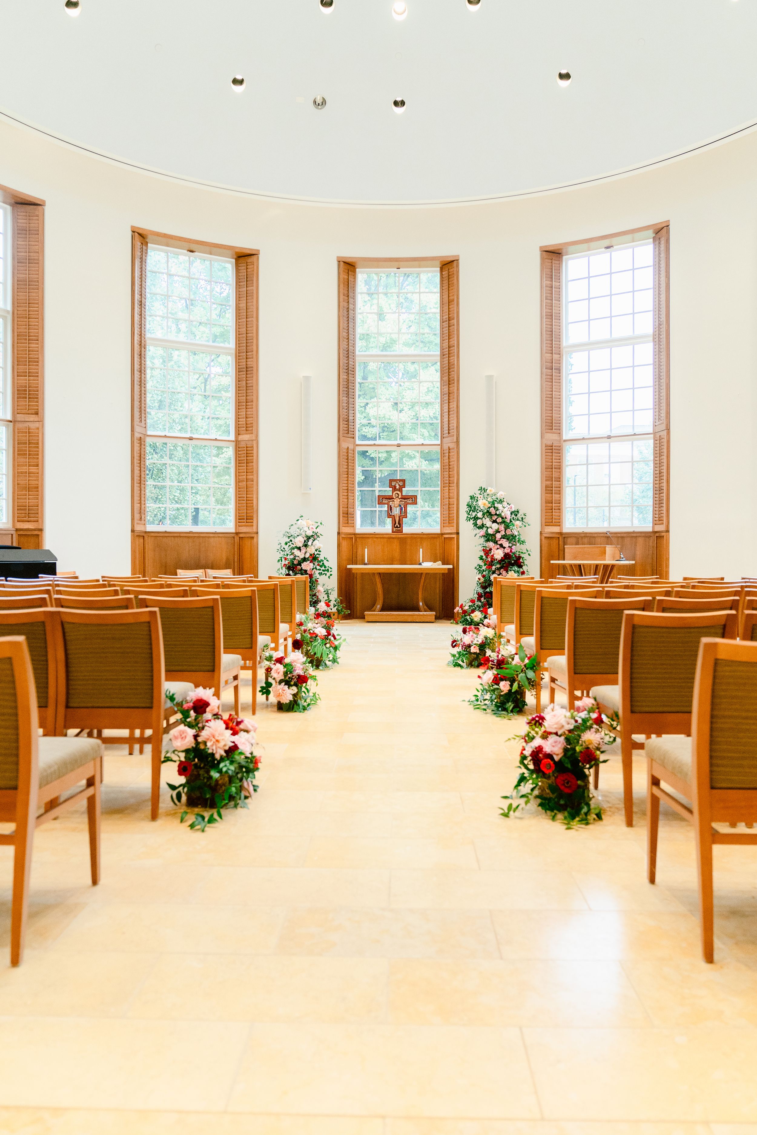 An elegant chapel interior with tall windows and floral arrangements lining the aisle for a wedding ceremony.