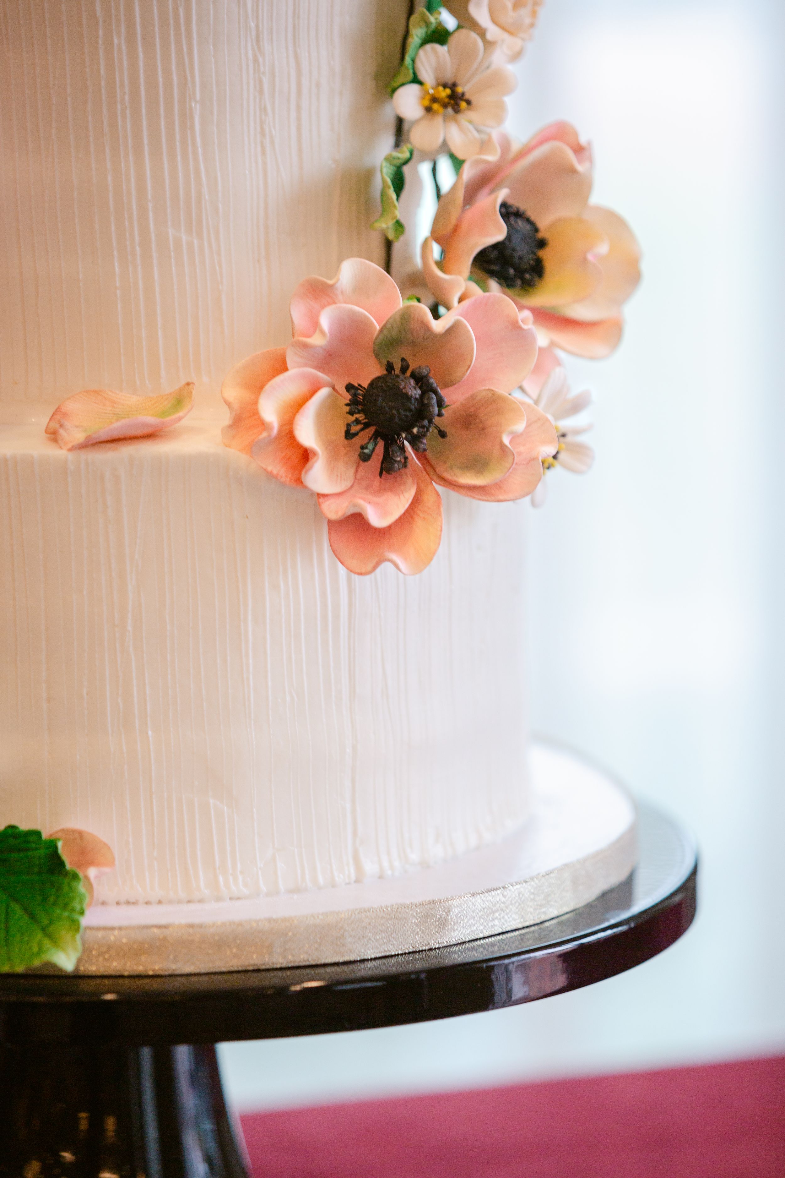 Close-up of elegant white wedding cake decorated with delicate peach-colored sugar flowers on dark cake stand.