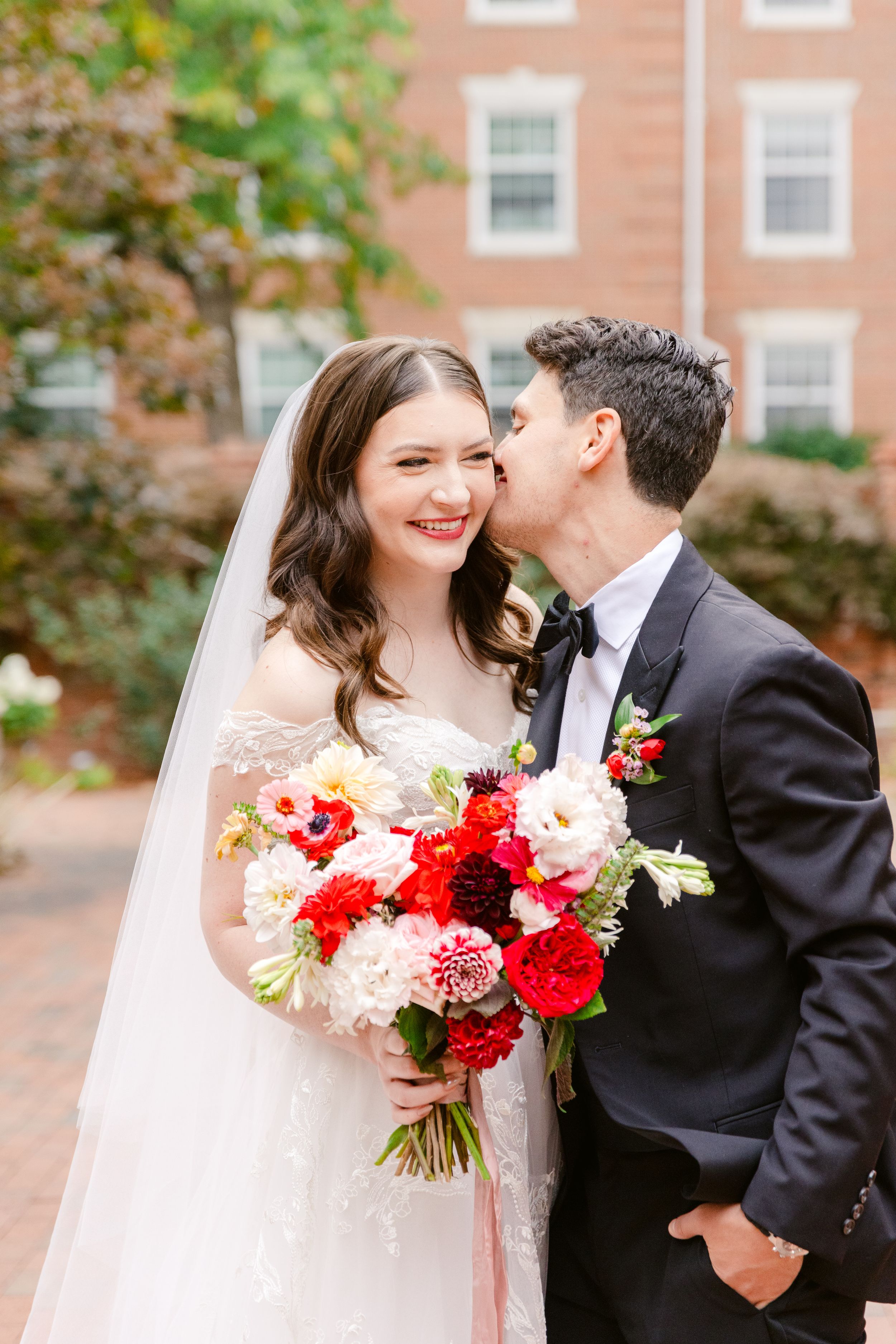 A couple in wedding attire share a tender moment outdoors with vibrant red and white bouquet against brick building.