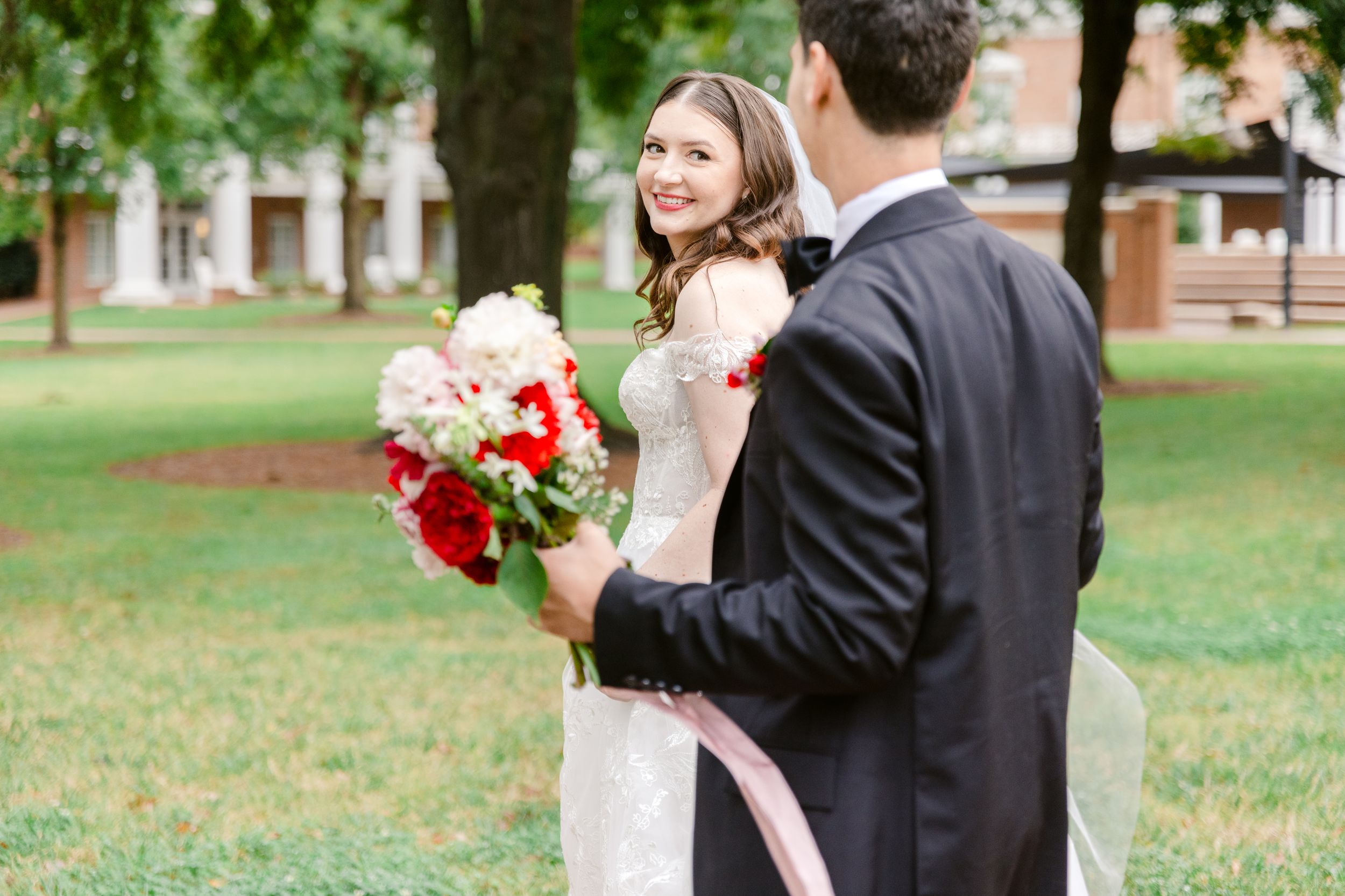 A couple in wedding attire shares a romantic moment outdoors on a grassy lawn with trees in the background.