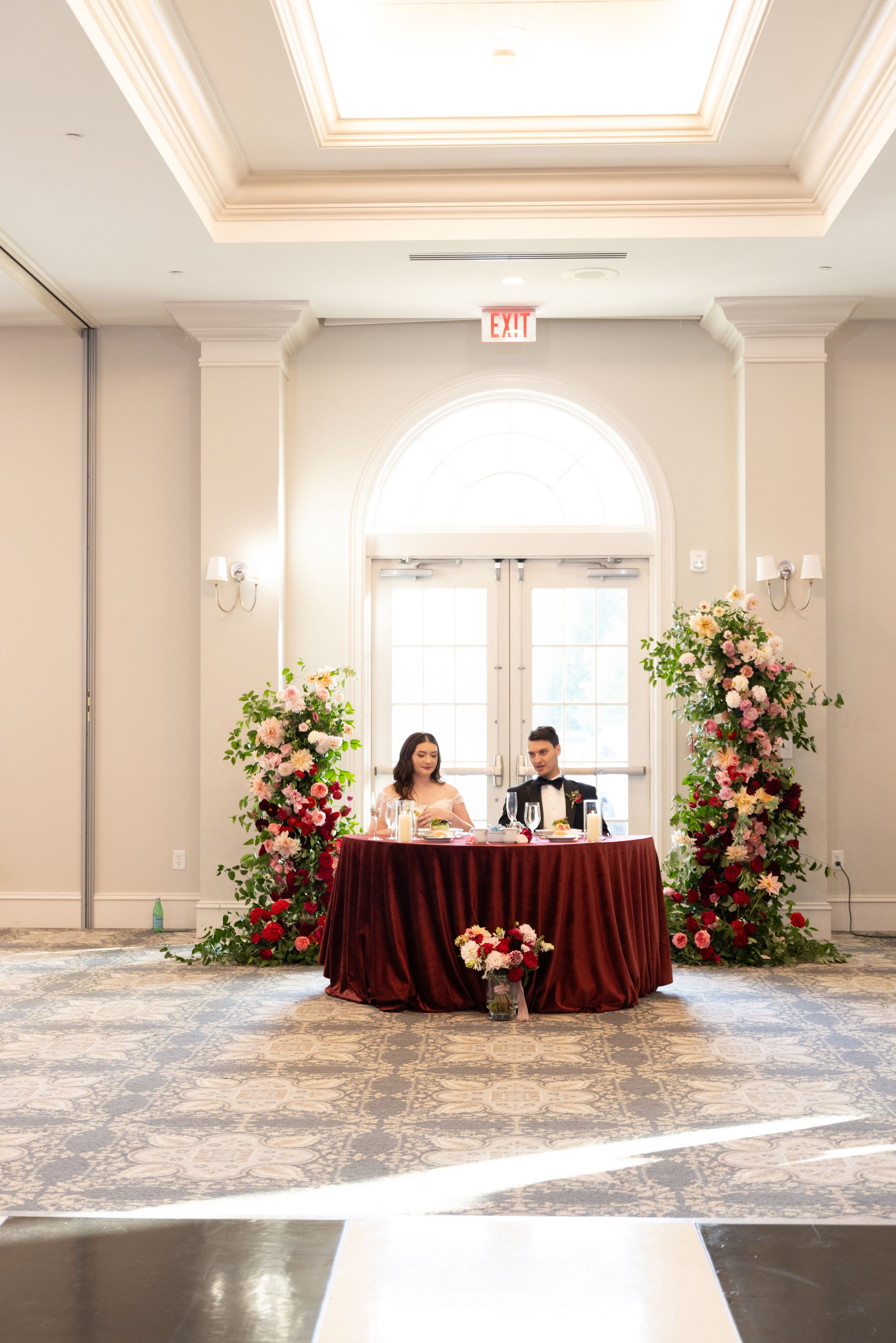 Elegant ballroom setup with burgundy tablecloths and pink floral arrangements along a light gray carpeted floor.