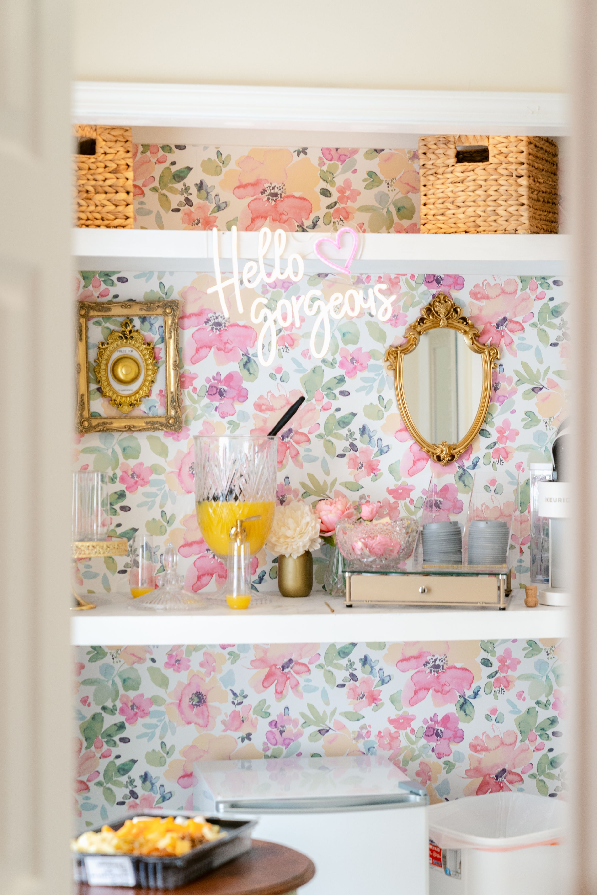 A romantic vanity area with floral wallpaper, gold mirrors, woven basket storage, and 'Hello Gorgeous' neon signage.