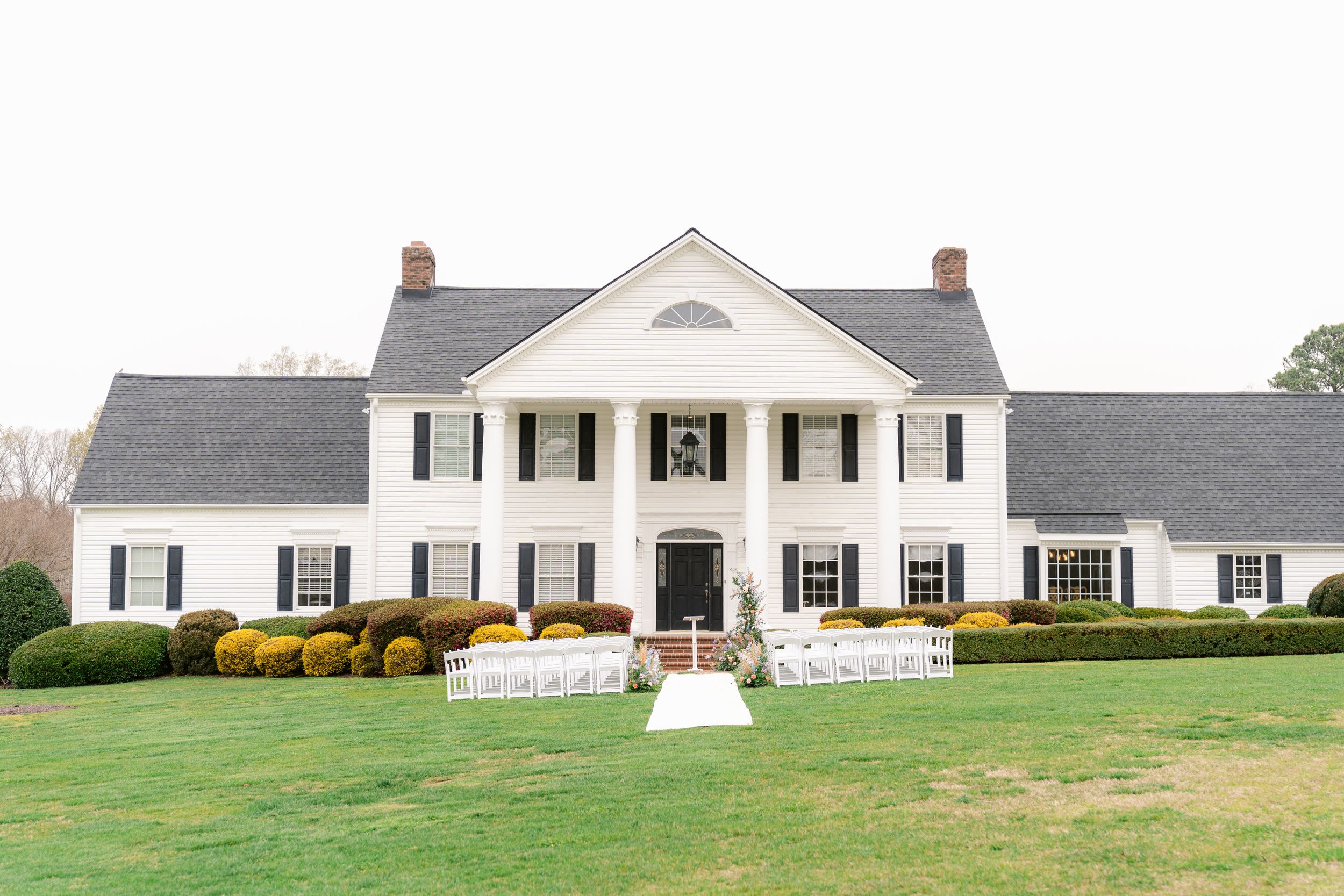 Elegant white colonial mansion with black shutters and yellow bushes sits on a manicured green lawn.