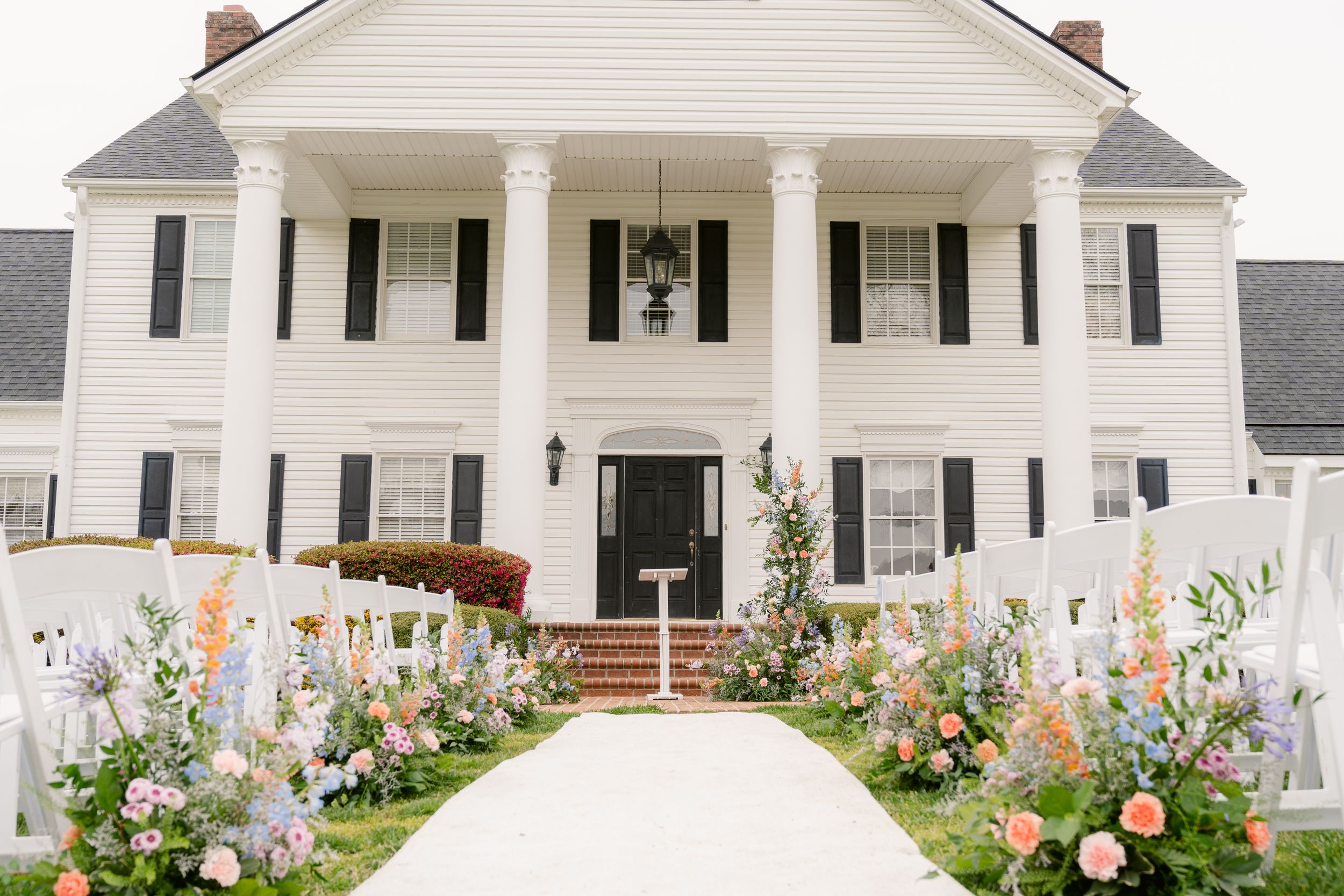 A white colonial mansion with grand columns features a symmetrical facade and colorful flower gardens lining the walkway.