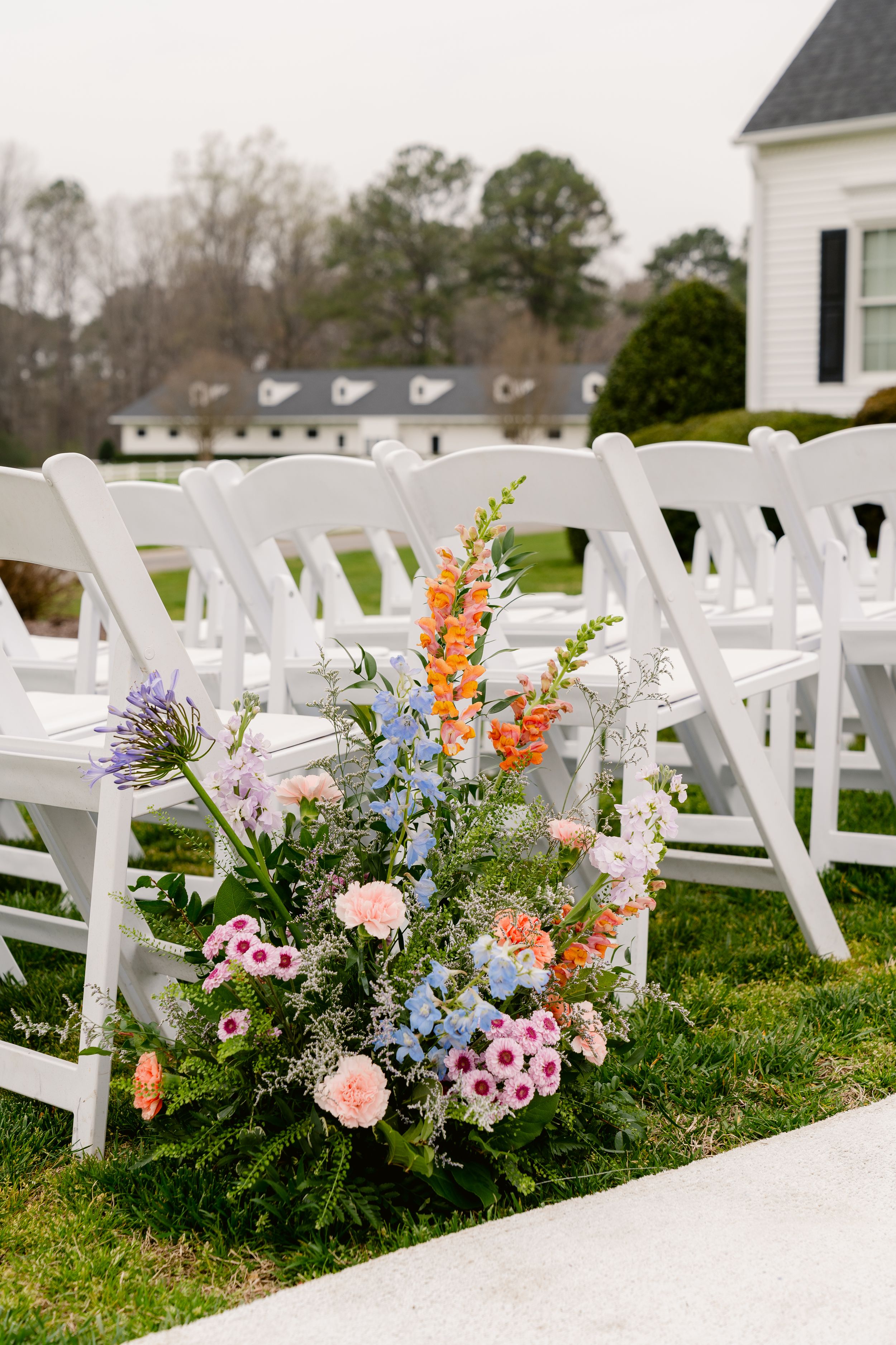Beautiful outdoor wedding ceremony setup with white chairs and colorful wildflower aisle arrangements in pink, orange and blue.