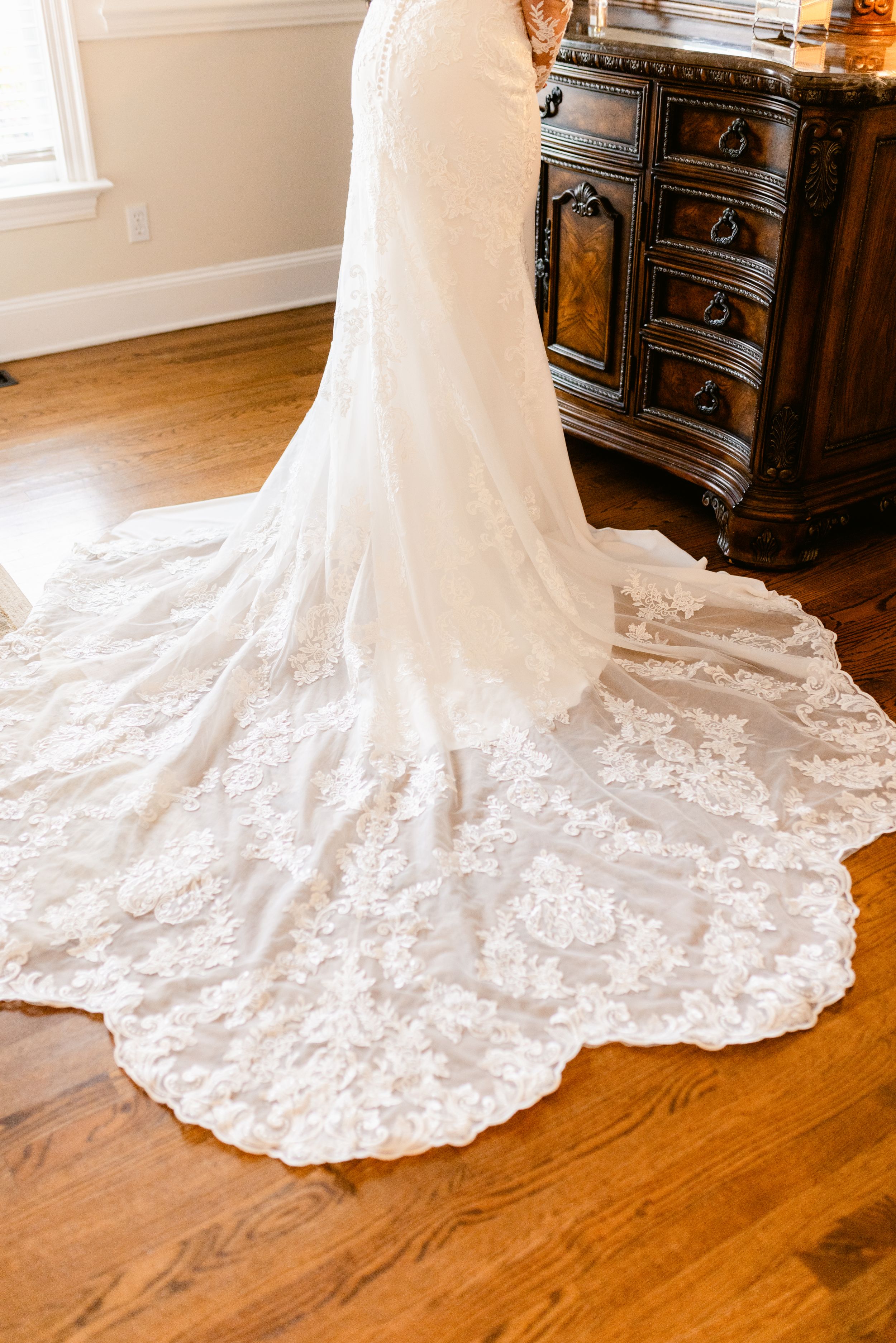 A stunning white lace wedding dress with a long cathedral train displayed on hardwood floors next to an ornate dresser.
