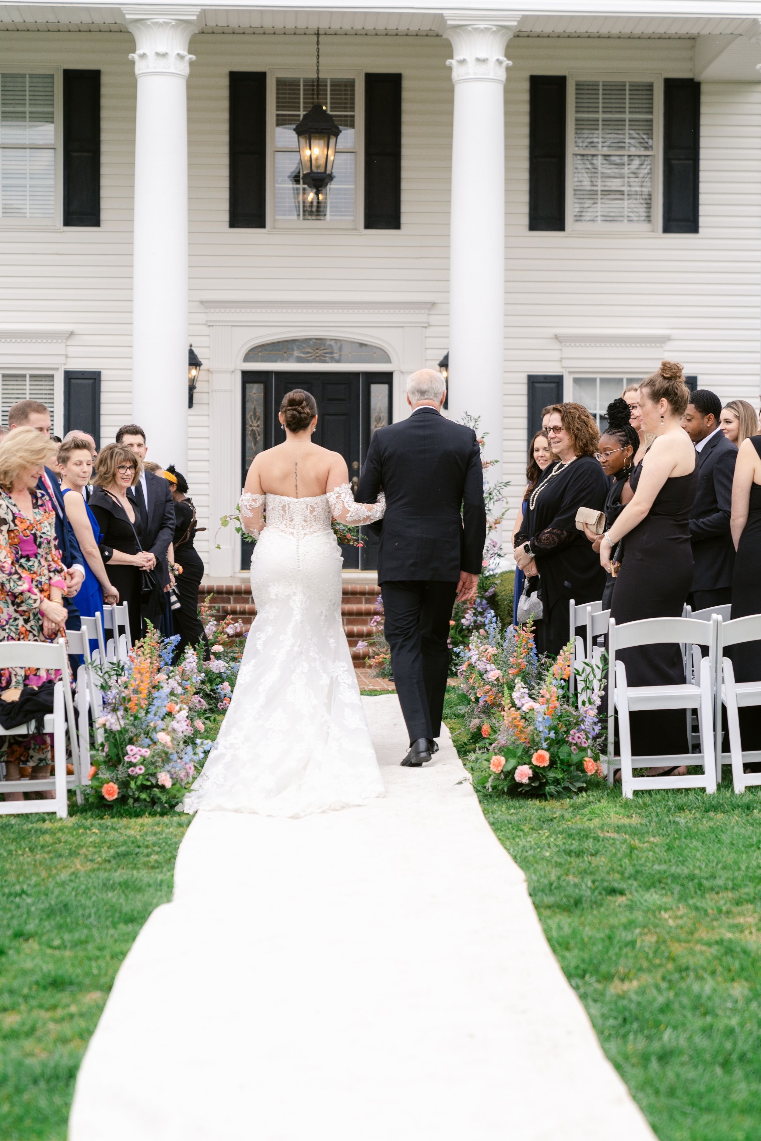 Wedding ceremony setup in front of a grand white colonial house with pillars and floral-lined aisle.