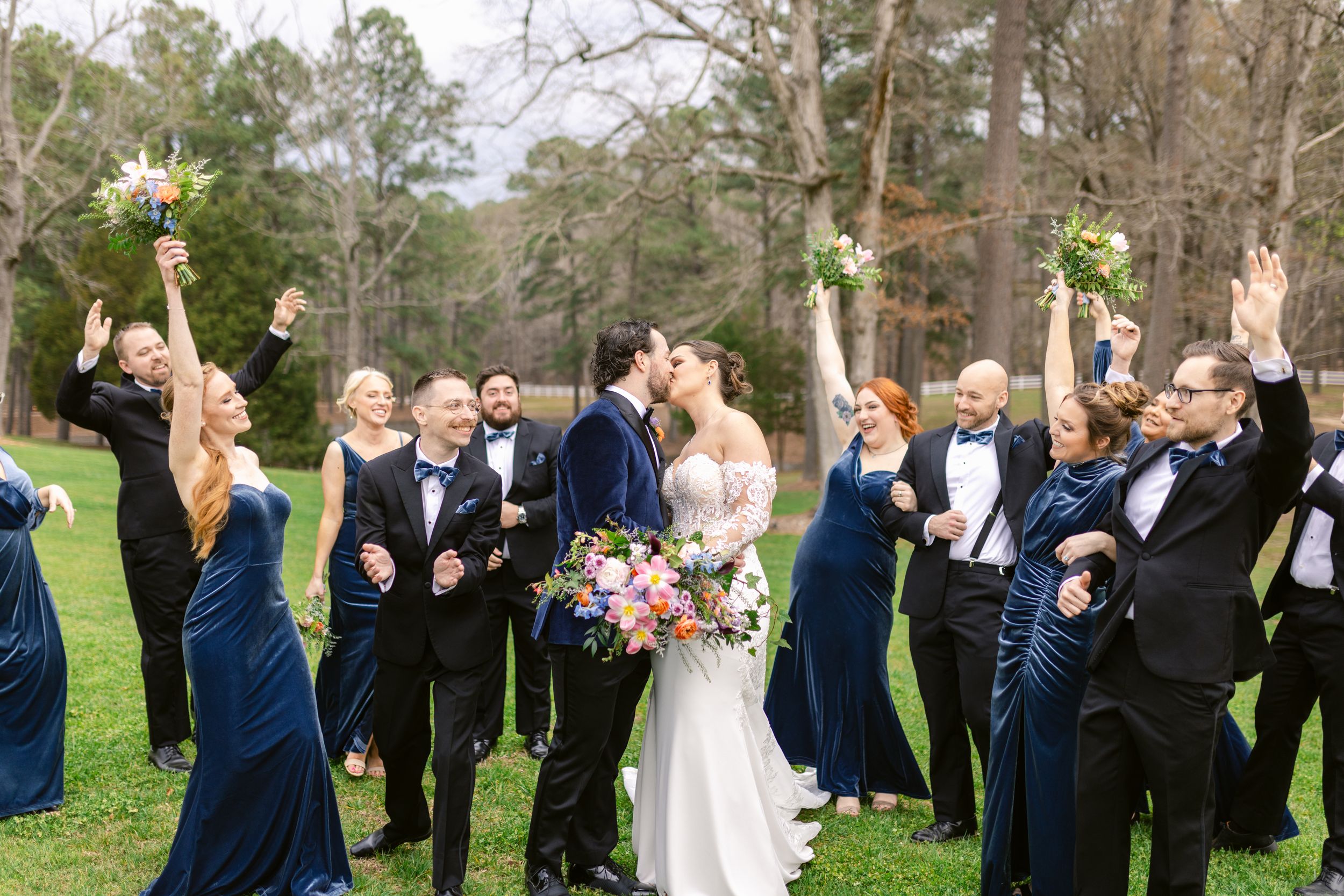 Wedding party in navy blue and white attire celebrating joyfully on a green lawn with trees in the background.