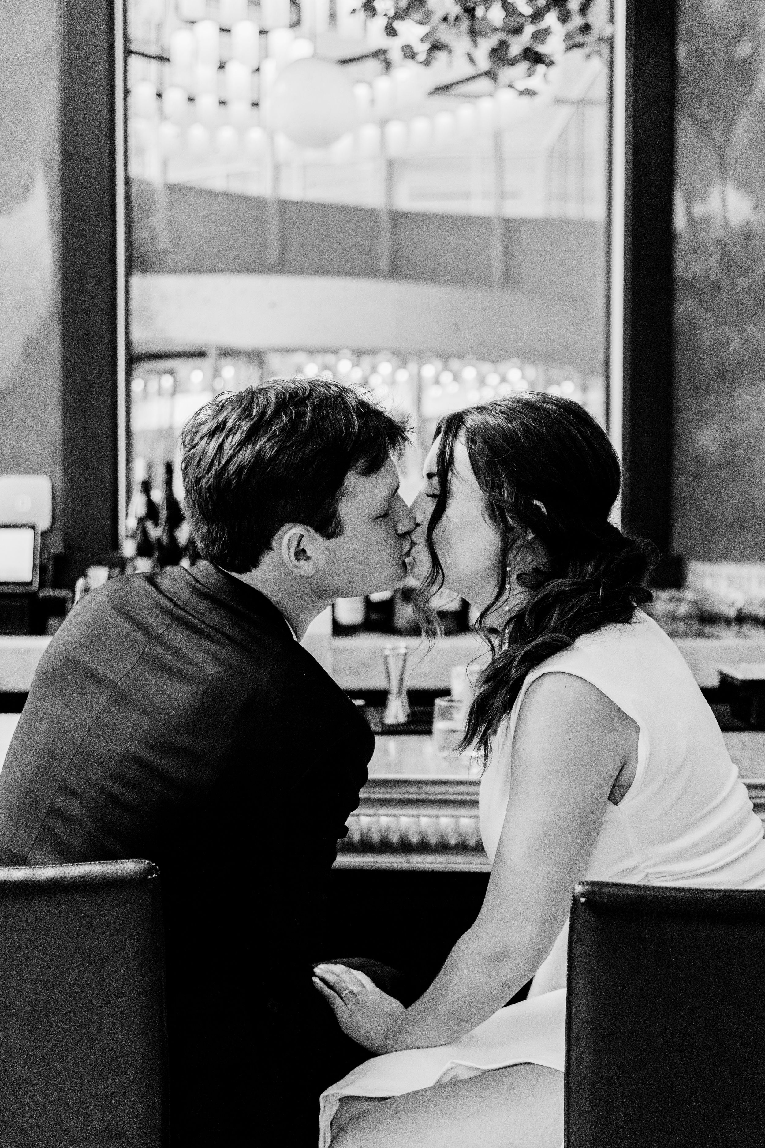 A romantic black and white photo of a couple kissing at a bar counter with hanging lights in the background.
