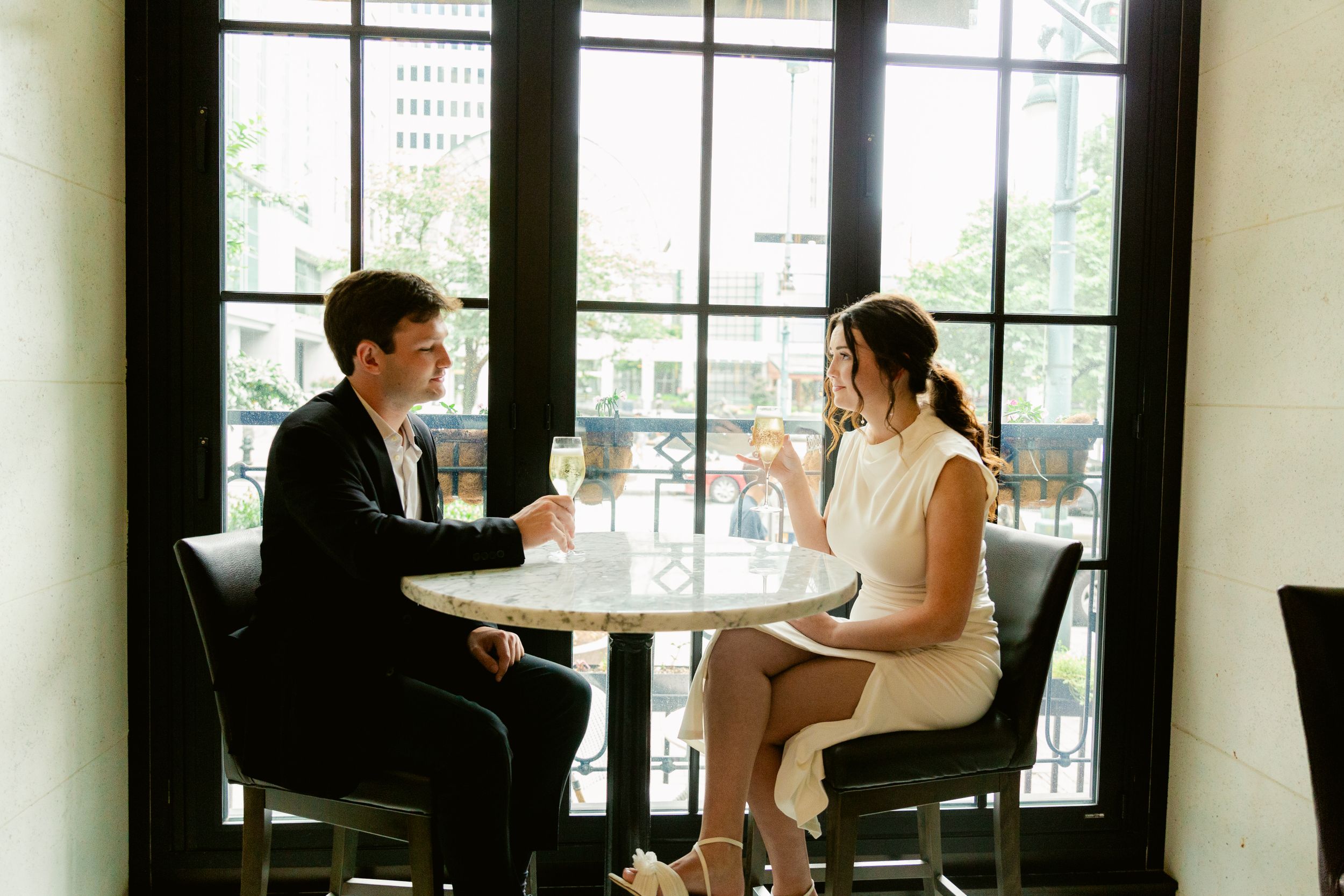 Two people having a professional conversation at a small cafe table near large windows in an elegant restaurant setting.