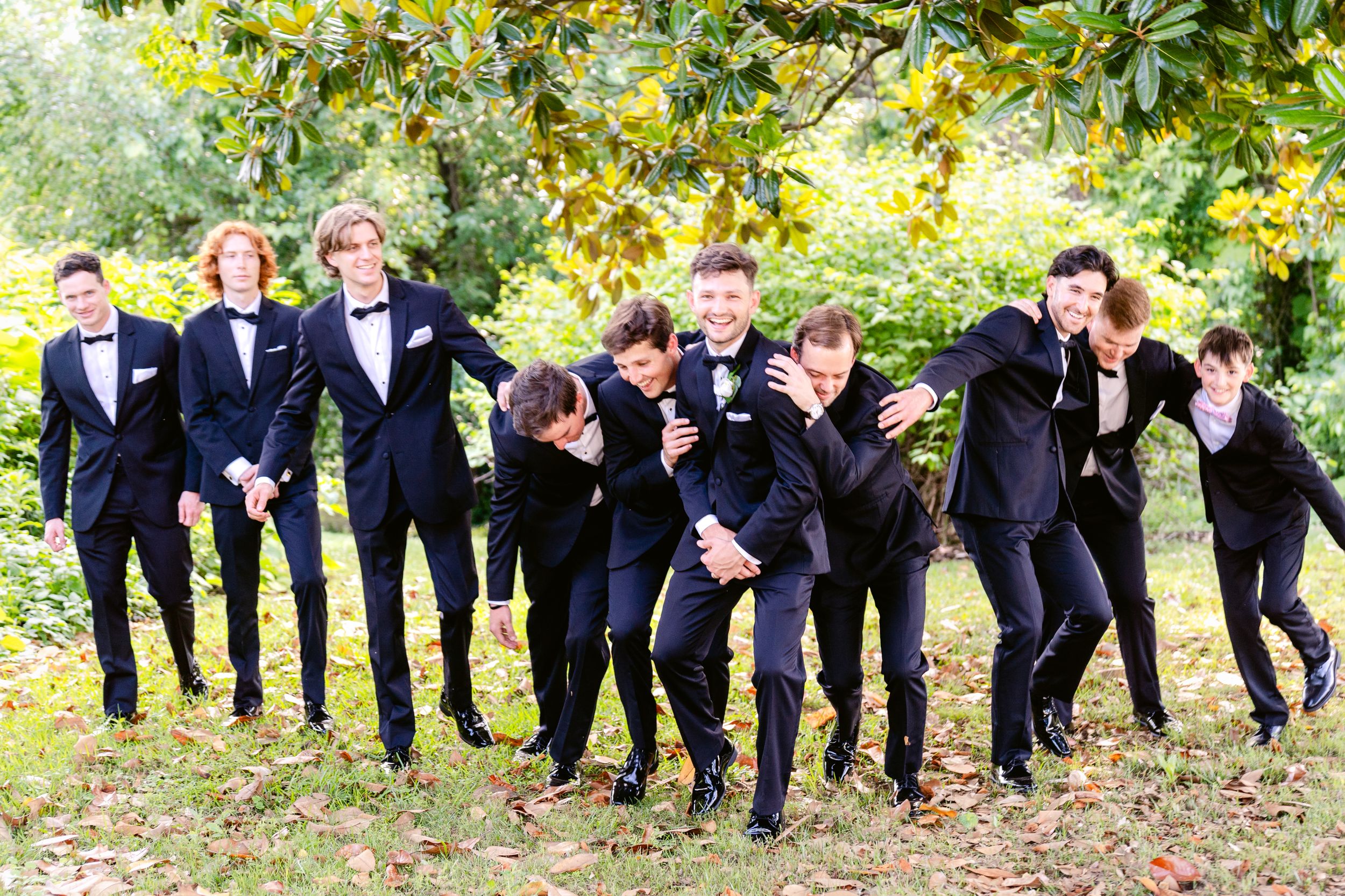 Wedding party in navy blue suits posing playfully and leaning to one side together outdoors under autumn trees.