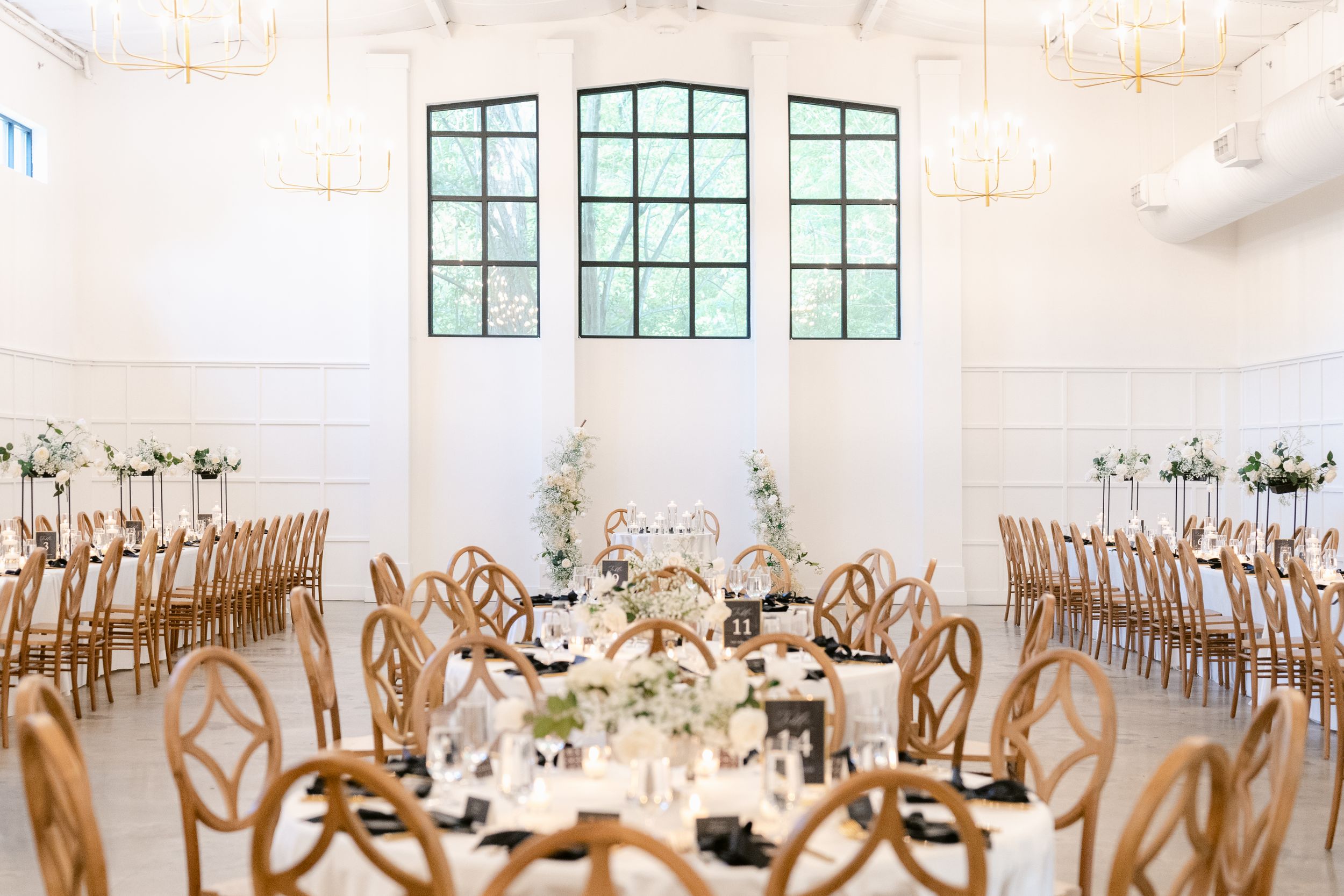 Elegant wedding reception venue with wooden chairs and white decor features tall windows and floral centerpieces.