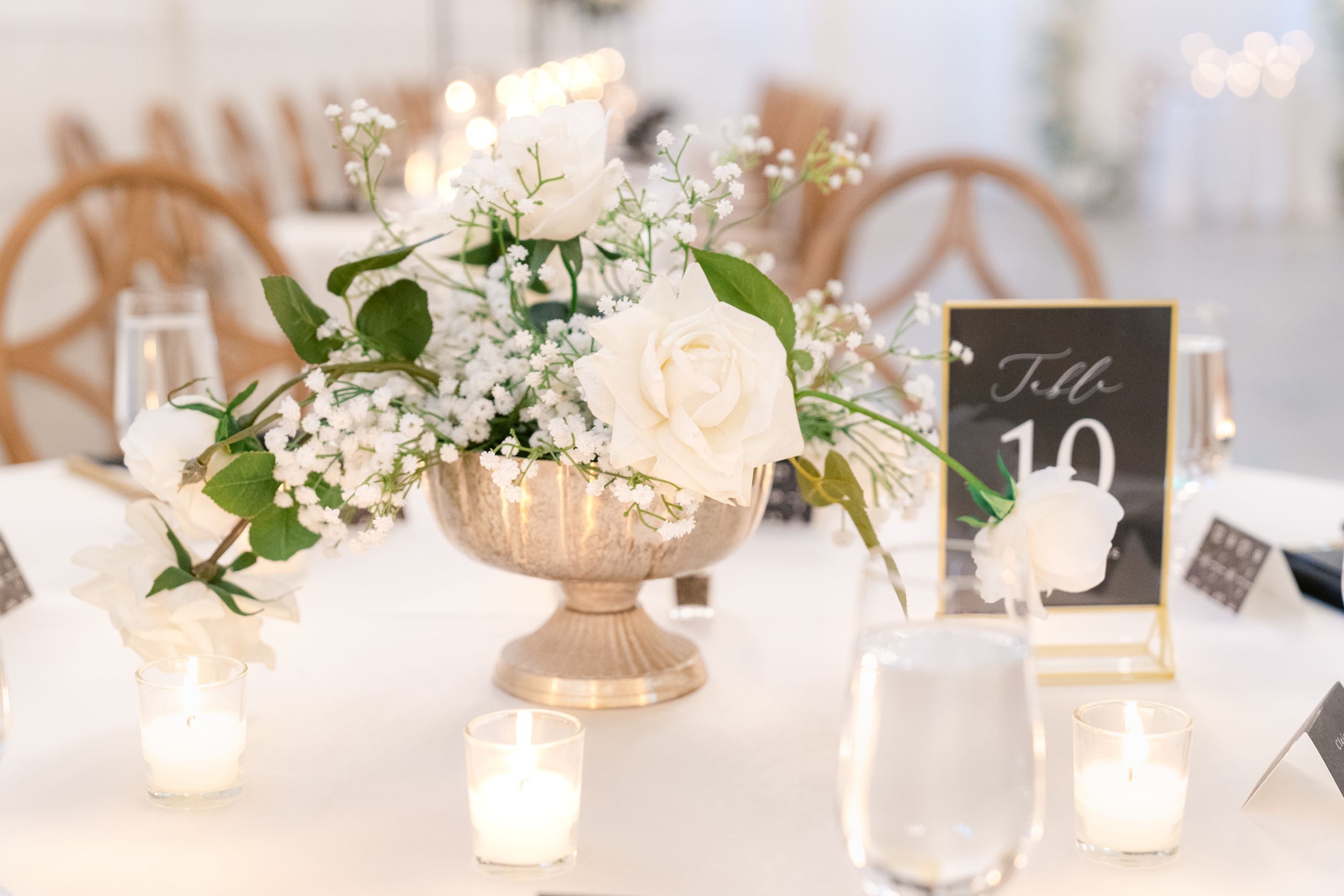 Elegant table setting with white roses and baby's breath in gold pedestal vase surrounded by glowing tea light candles.