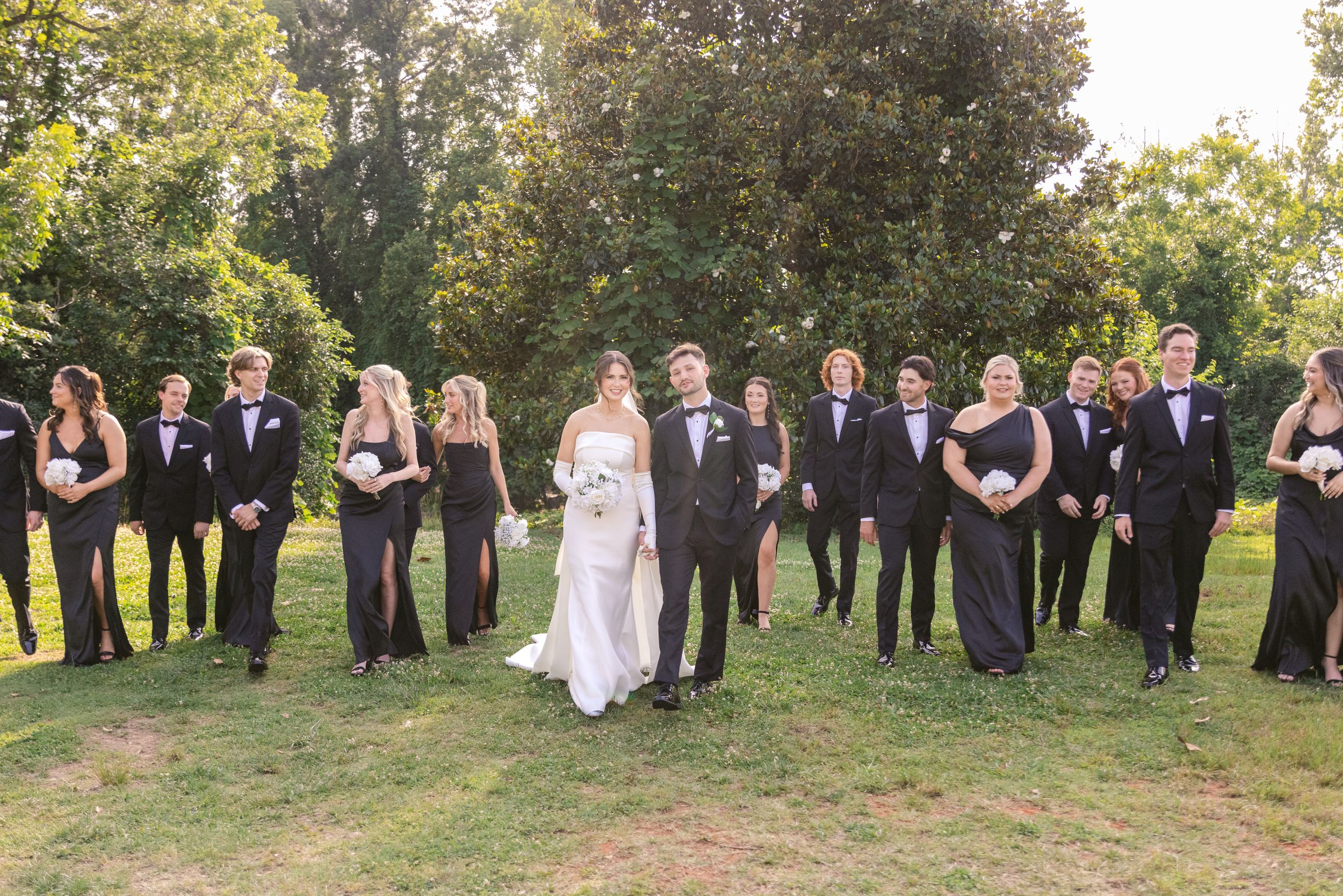 Wedding party in formal attire poses on grass with trees in background during outdoor photo session.