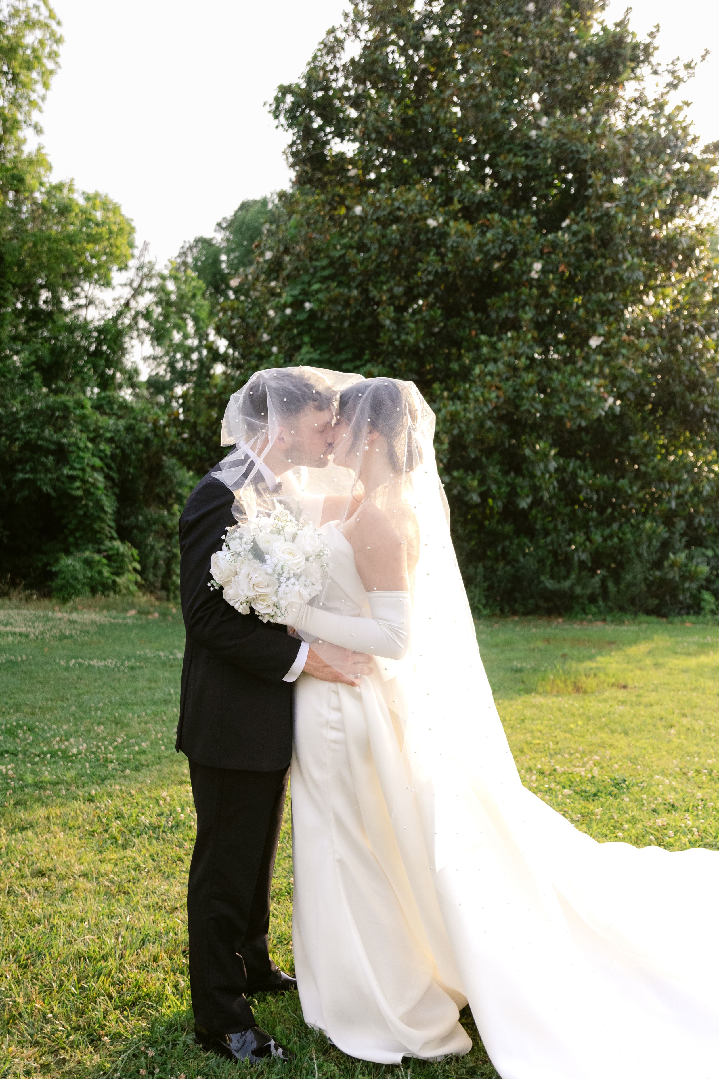 A bride and groom share an intimate moment under a veil in a garden setting during golden hour.