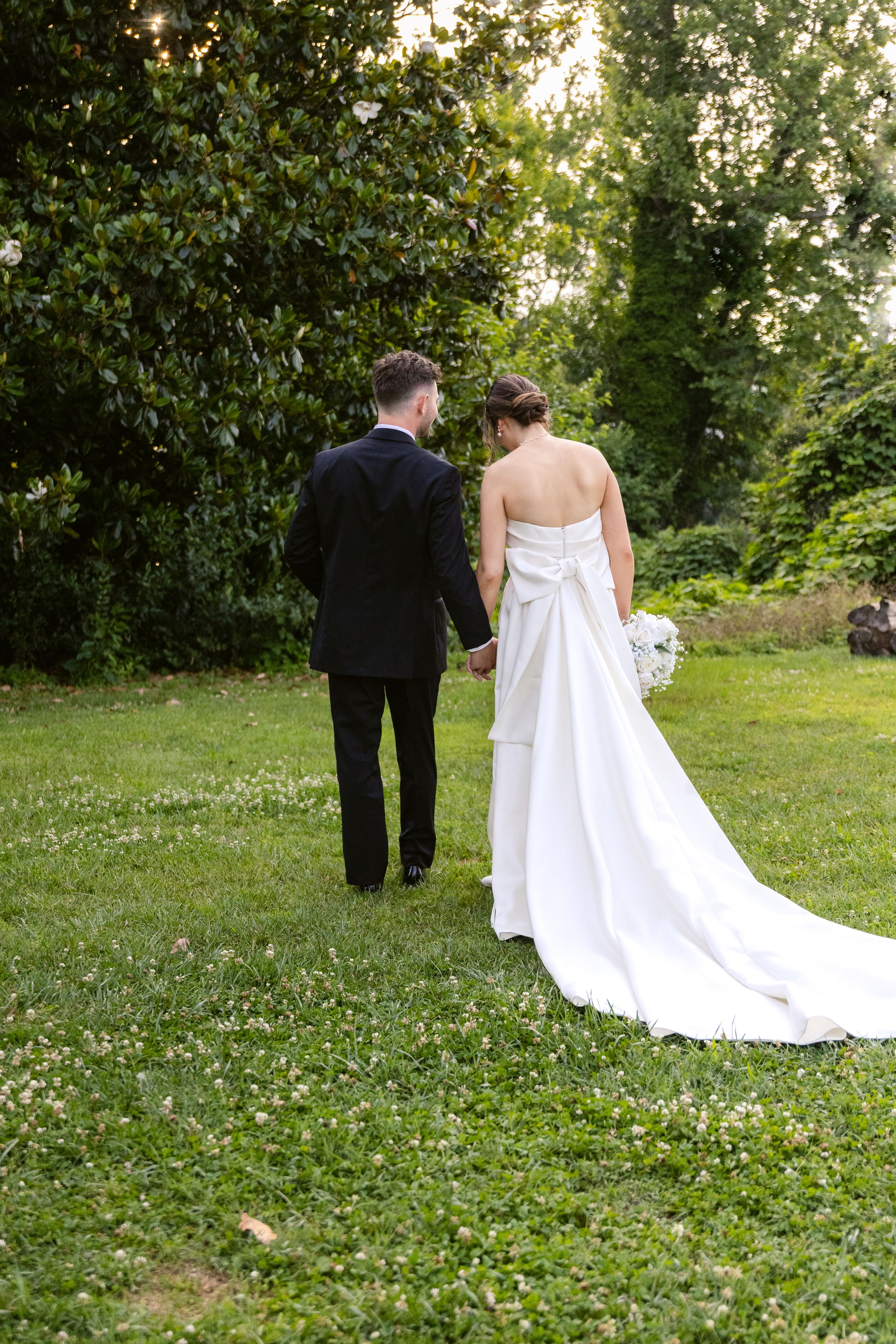 A newlywed couple walks together on a lush green lawn surrounded by trees, the bride wearing a flowing white gown.