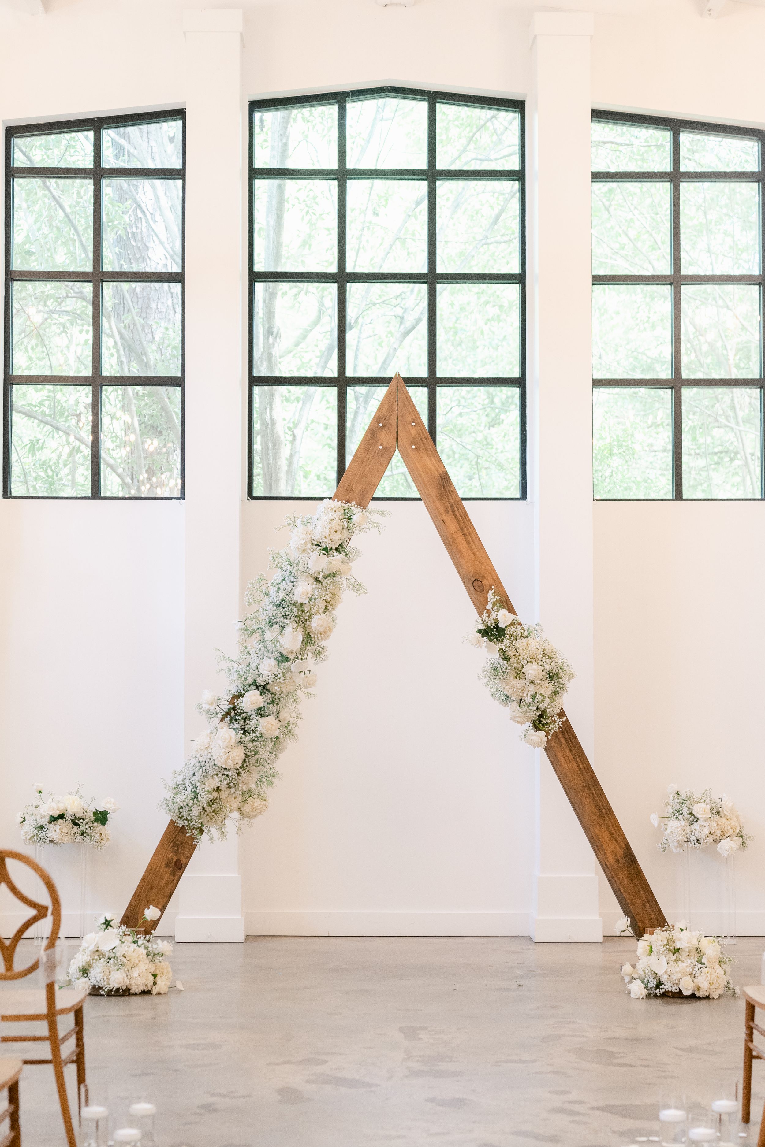 Modern wedding ceremony setup with wooden triangle arches and white floral arrangements in an industrial space.