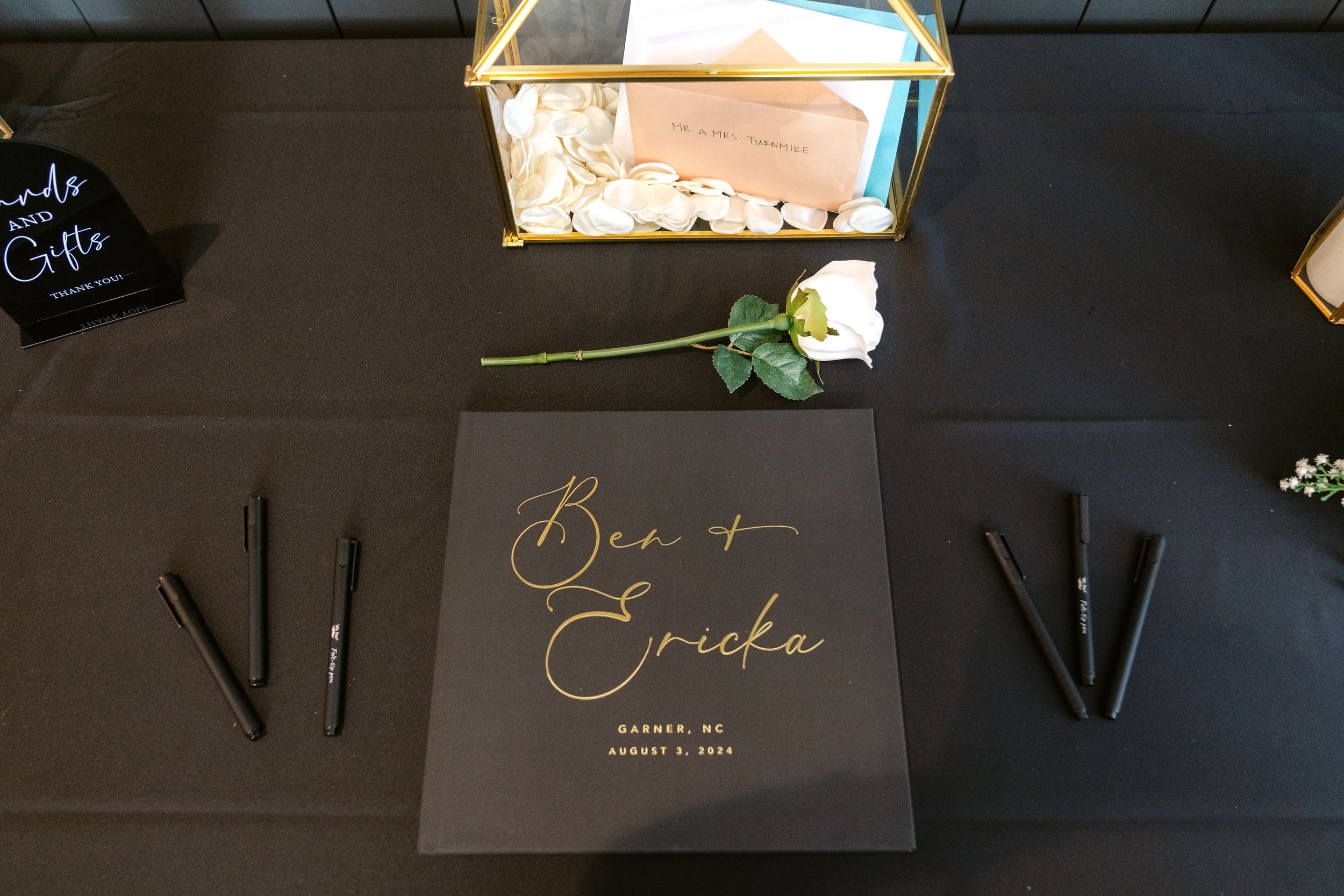 Black and gold wedding guest book with calligraphy names and pens on dark table with rose accent.