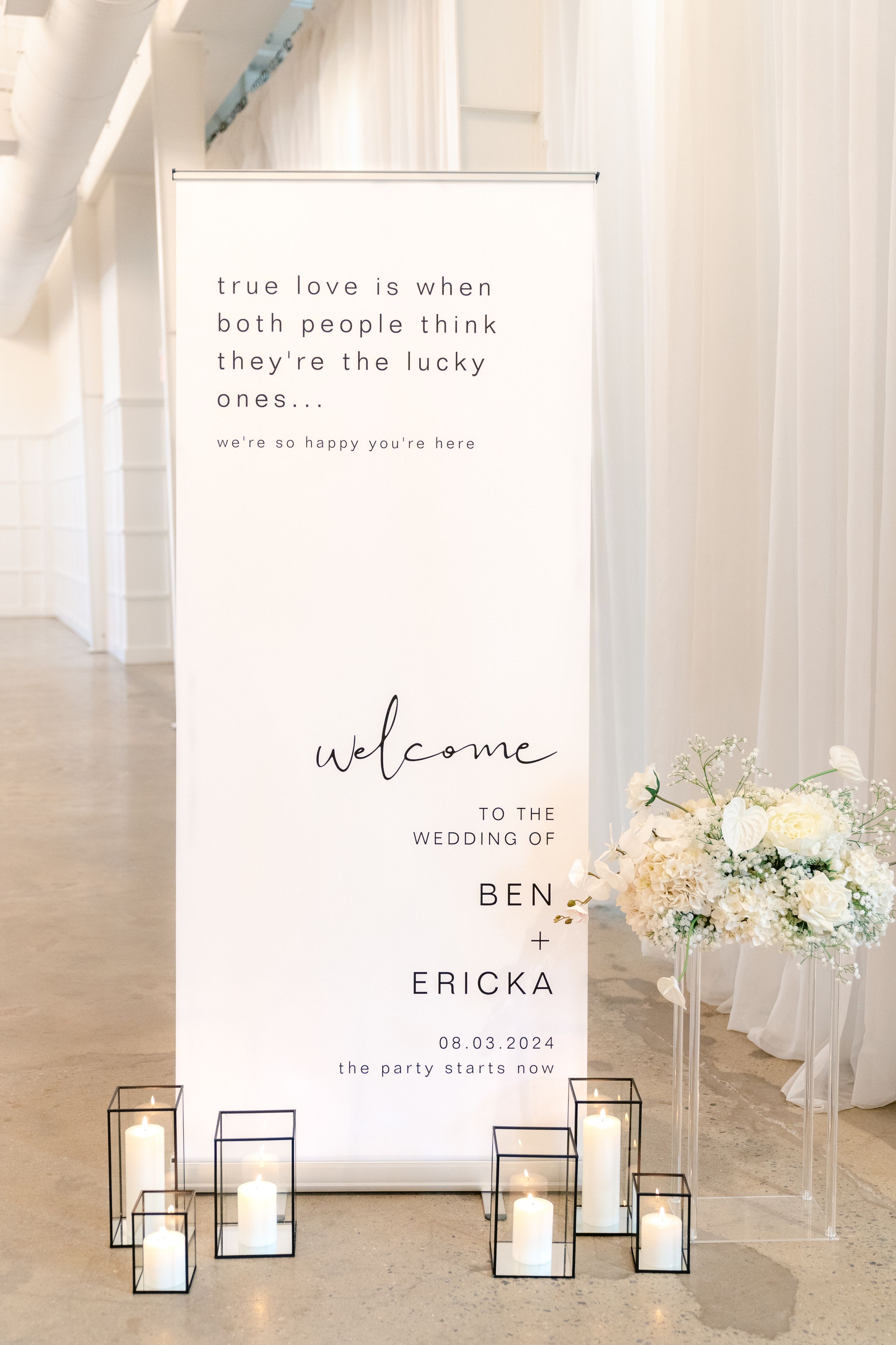 Minimalist wedding welcome sign display with candles and floral arrangements in a bright white venue.