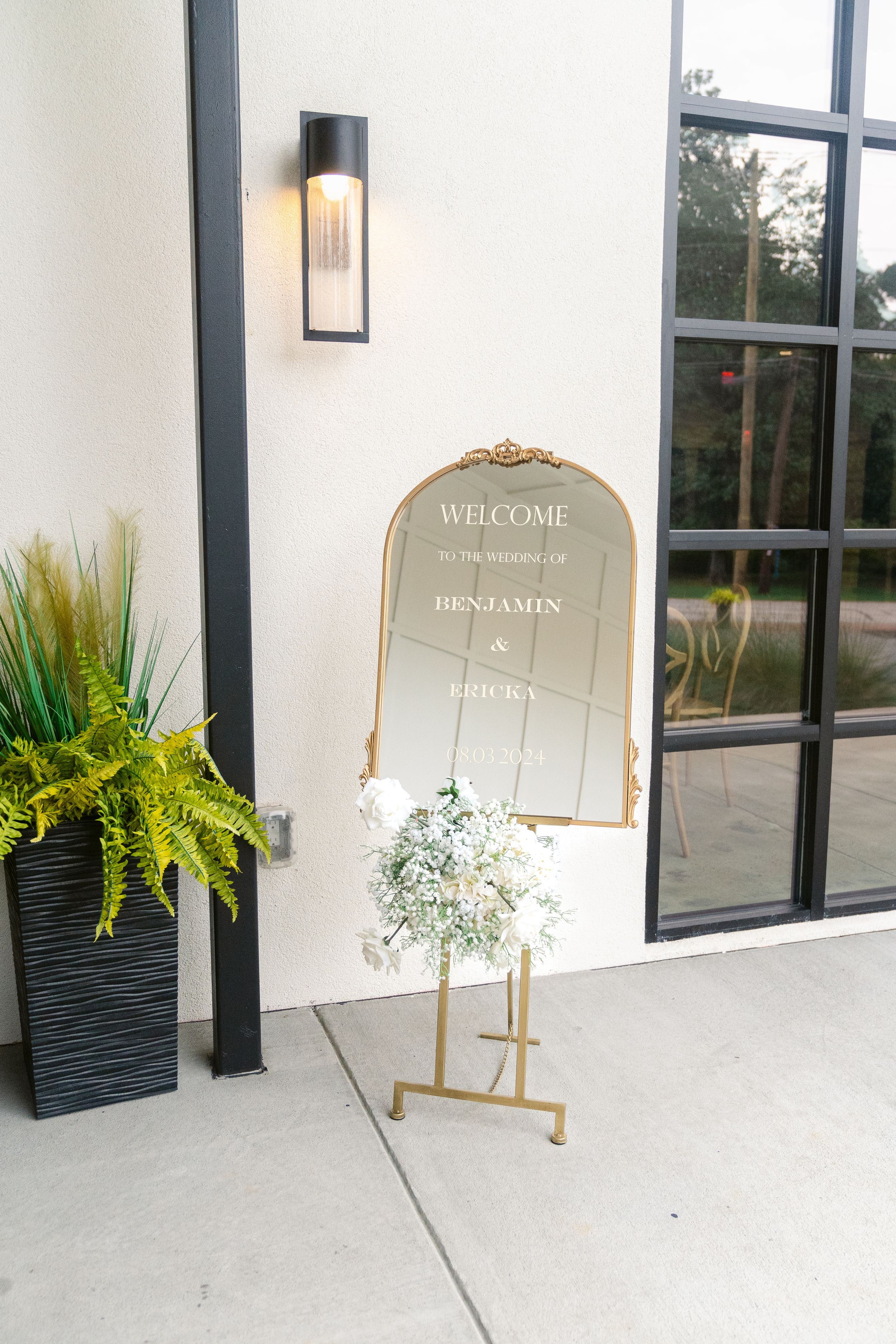 Elegant gold-framed welcome mirror sign with potted fern decoration outside venue entrance.
