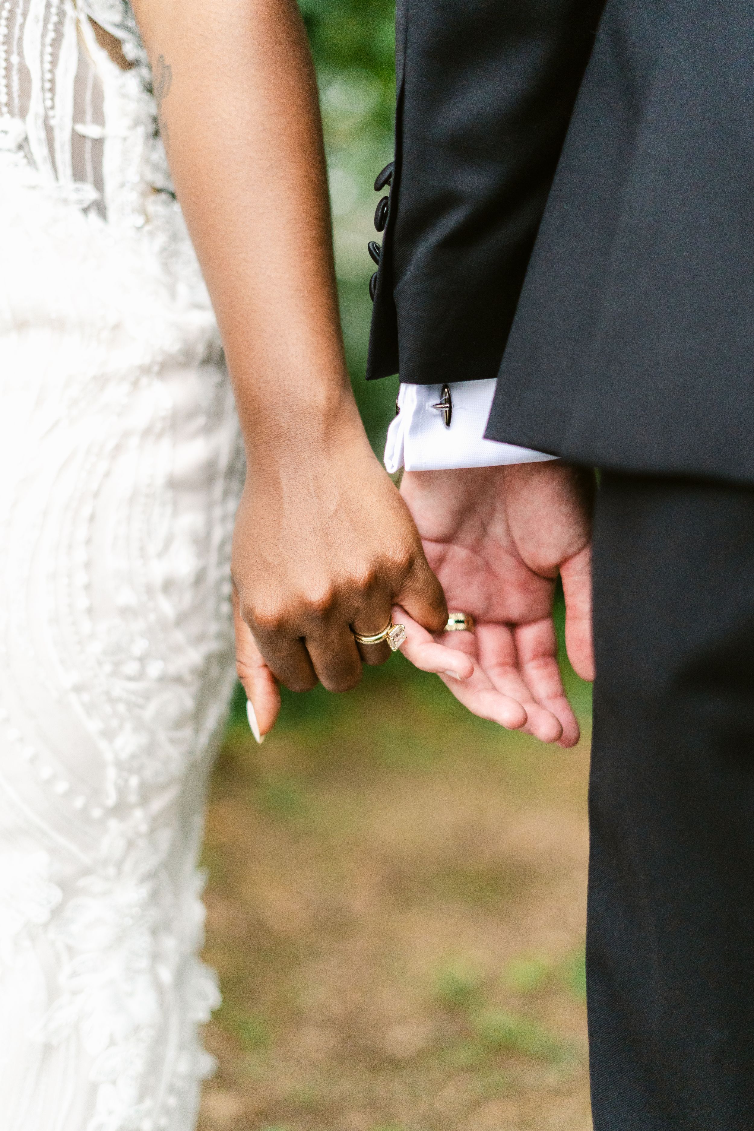 Close-up of newlyweds holding hands, showing white lace dress and black suit details.