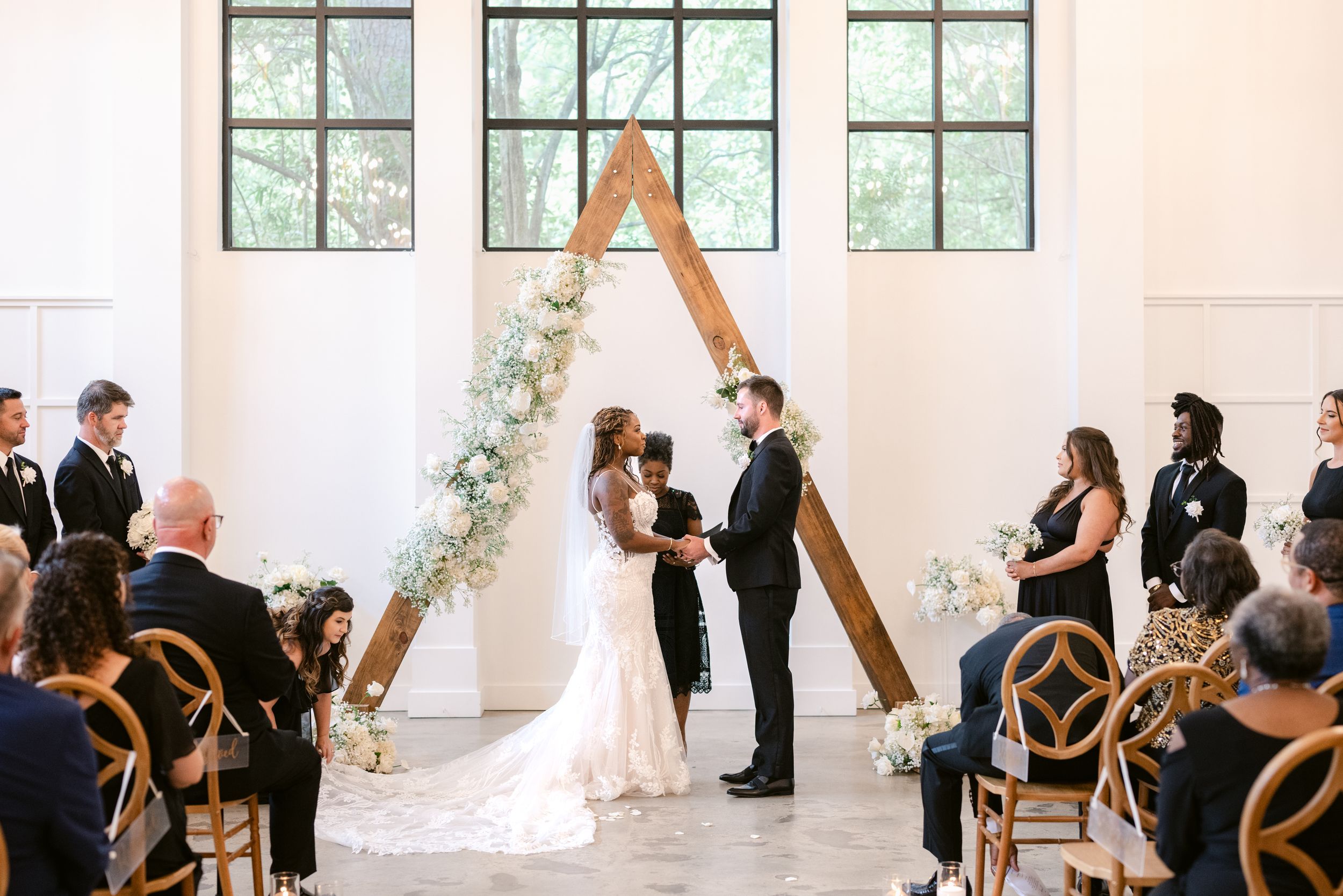 A wedding ceremony takes place in a bright white room with large windows and wooden cross-back chairs.