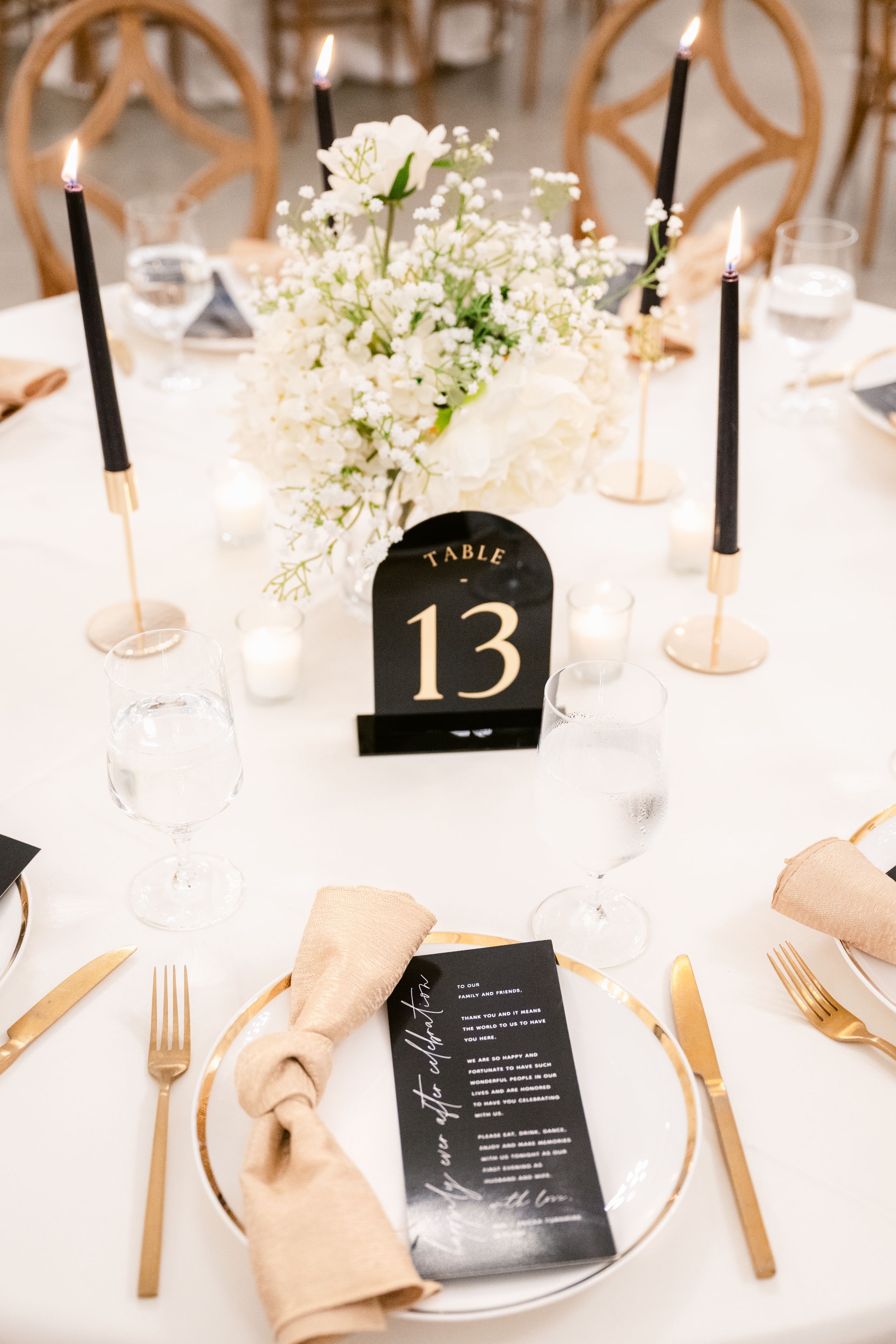 Elegant wedding table setting with black menu cards, gold cutlery, and white floral centerpieces against a crisp white tablecloth.