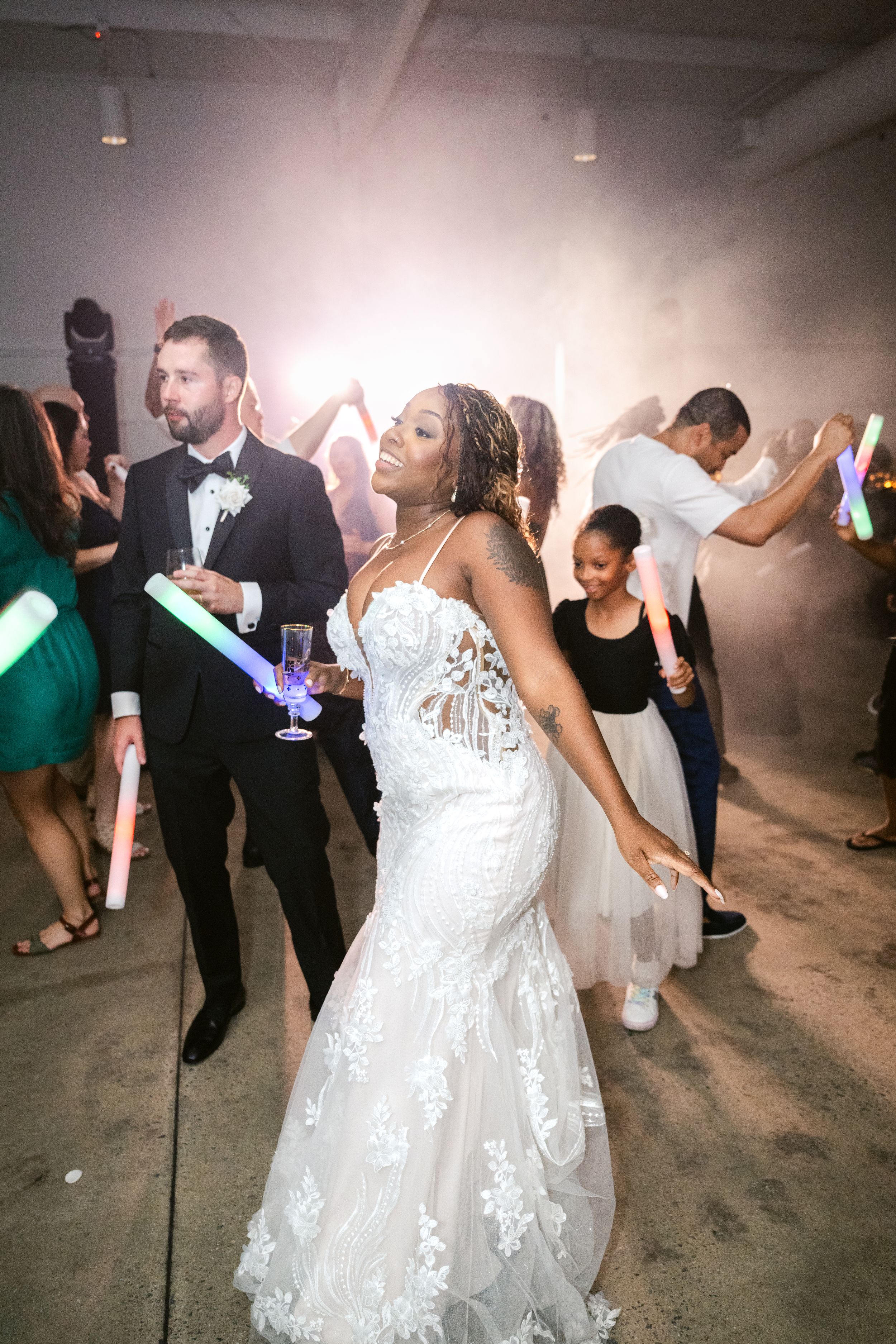 A person in a white lace wedding dress dances joyfully at a reception celebration with other guests on the dance floor.