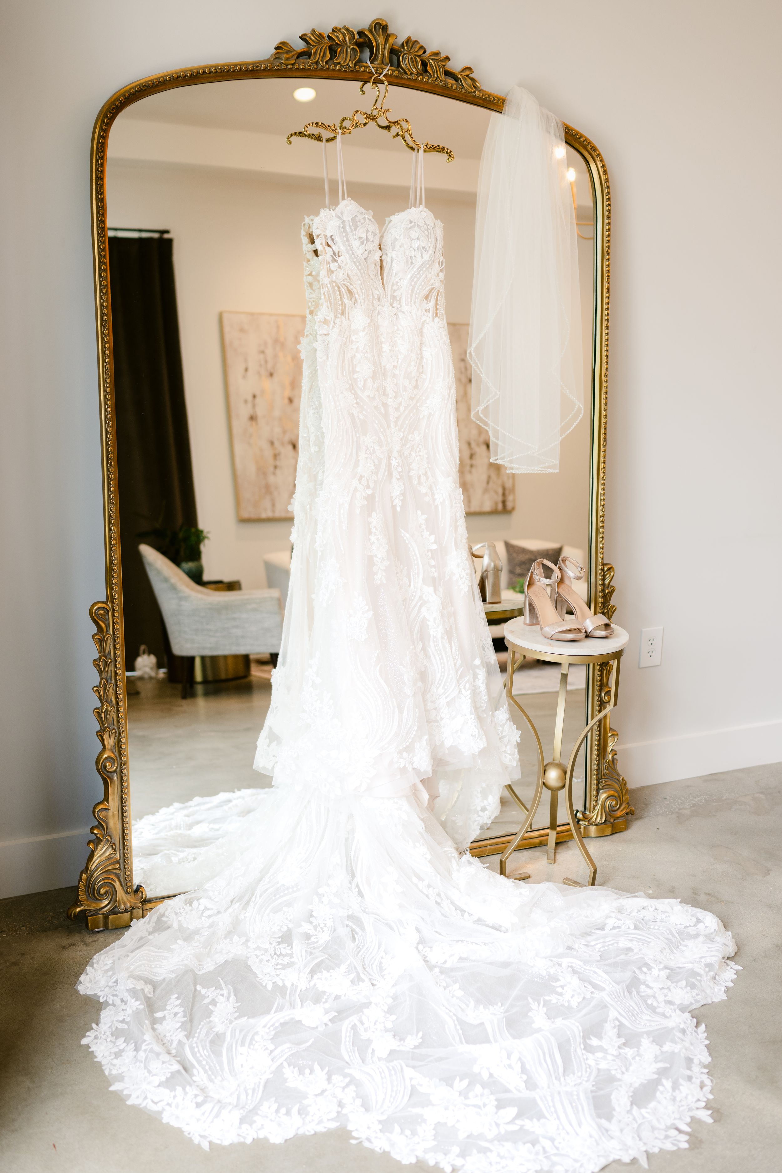 Stunning lace wedding dress with long train displayed on hanger in front of ornate gold mirror.