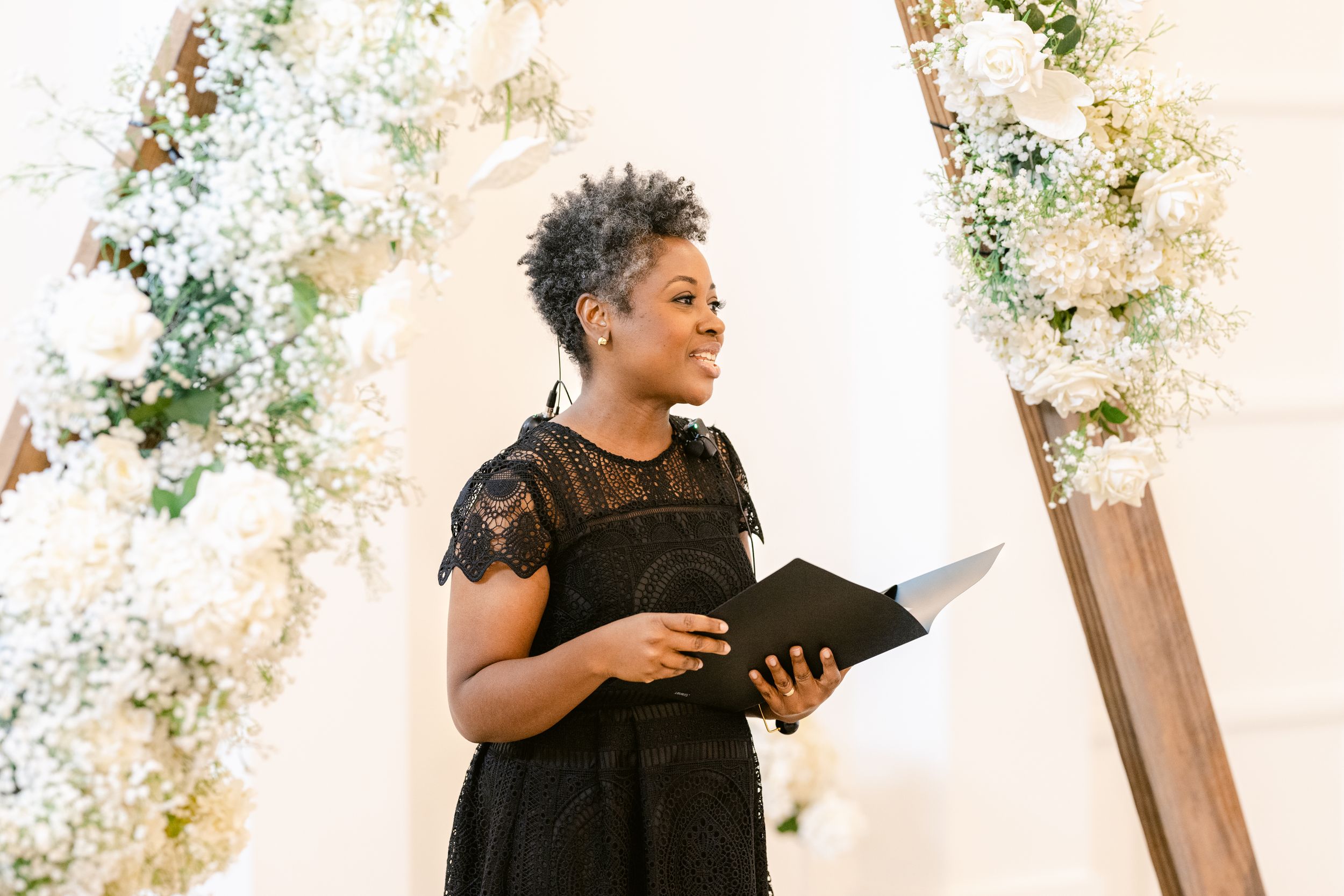 Wedding officiant in black lace dress reads from book at altar decorated with white floral arrangements.