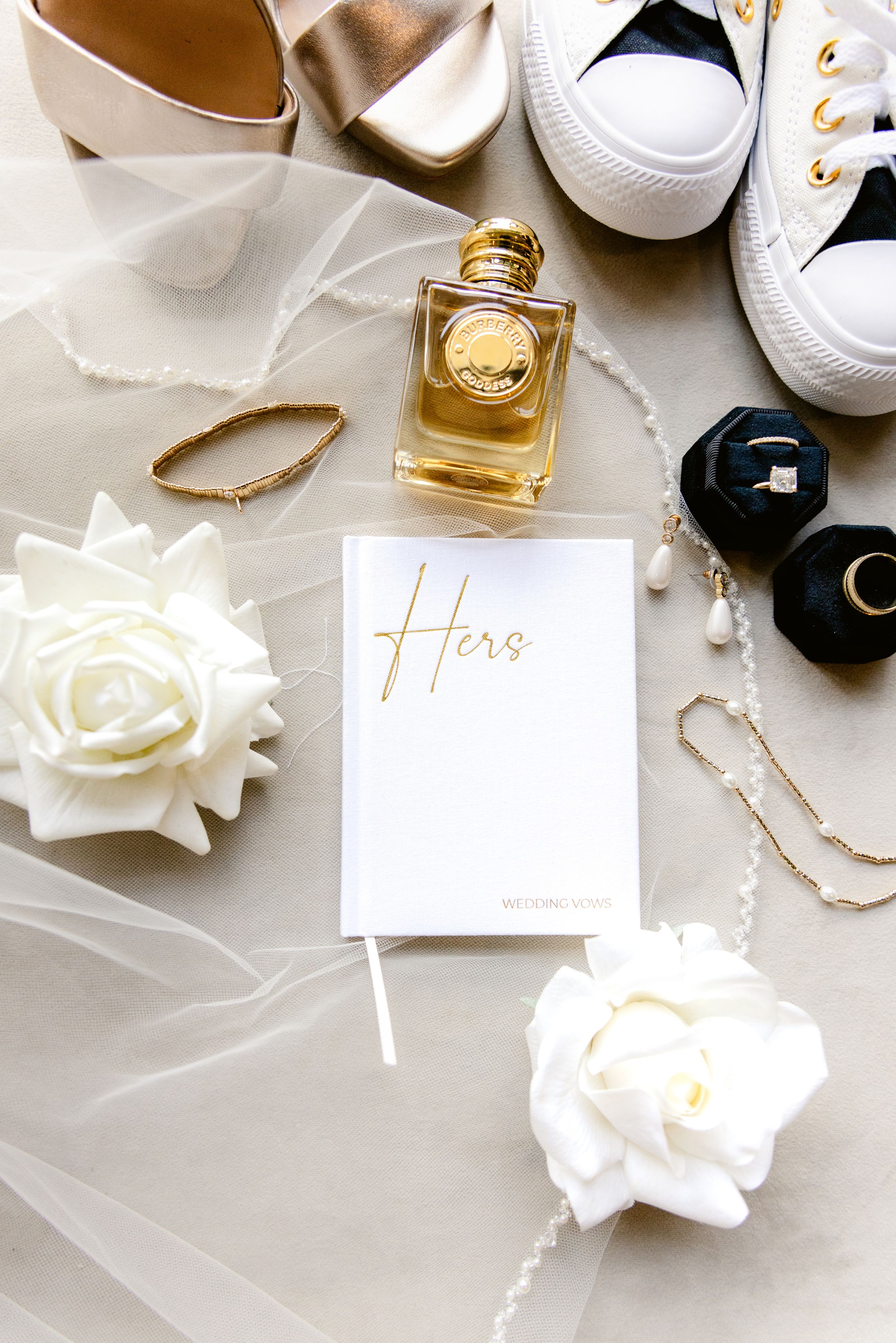 Elegant wedding details including white roses, perfume bottle, jewelry and shoes arranged on light fabric.