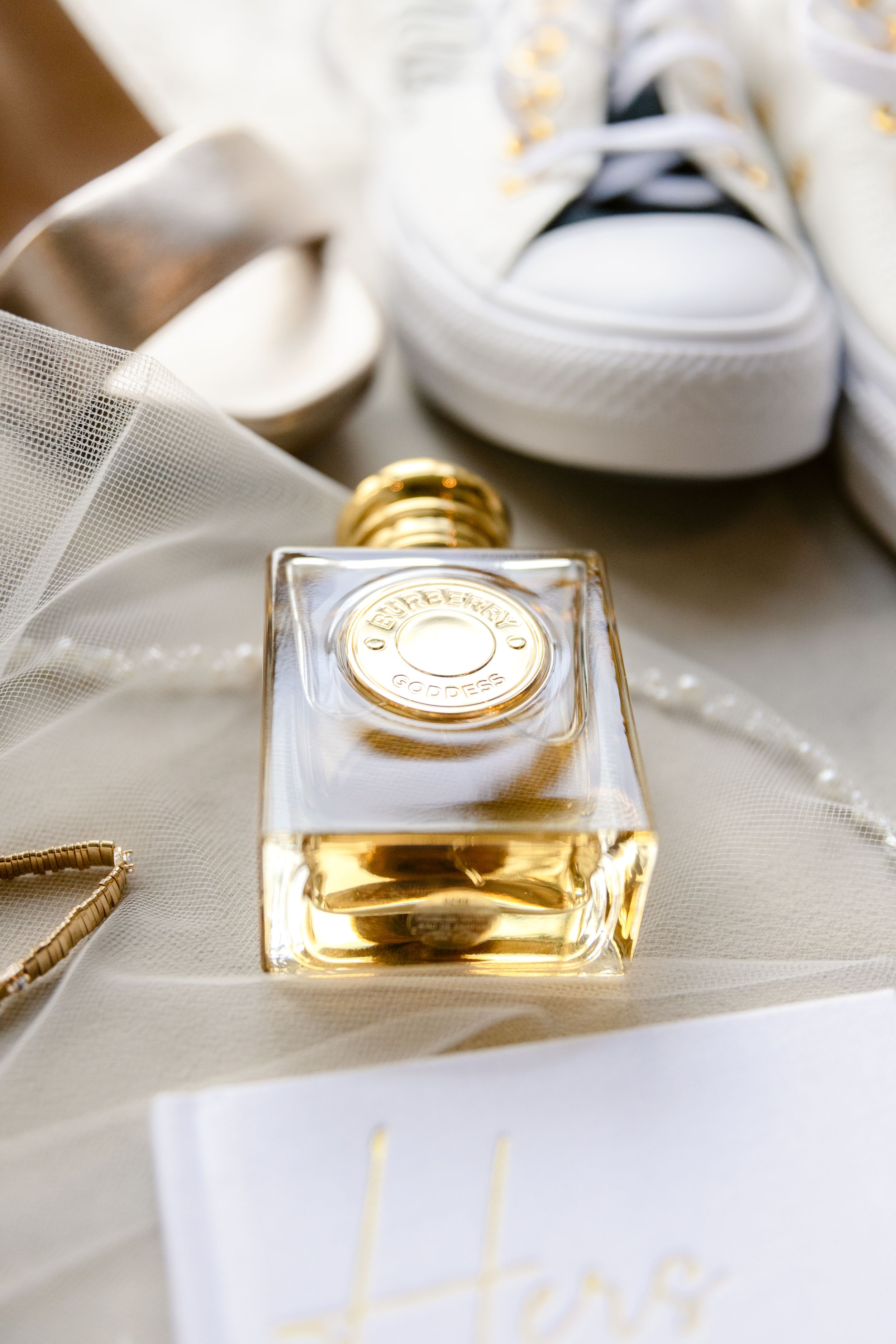 Elegant perfume bottle with gold cap displayed next to white sneakers and delicate tulle fabric.