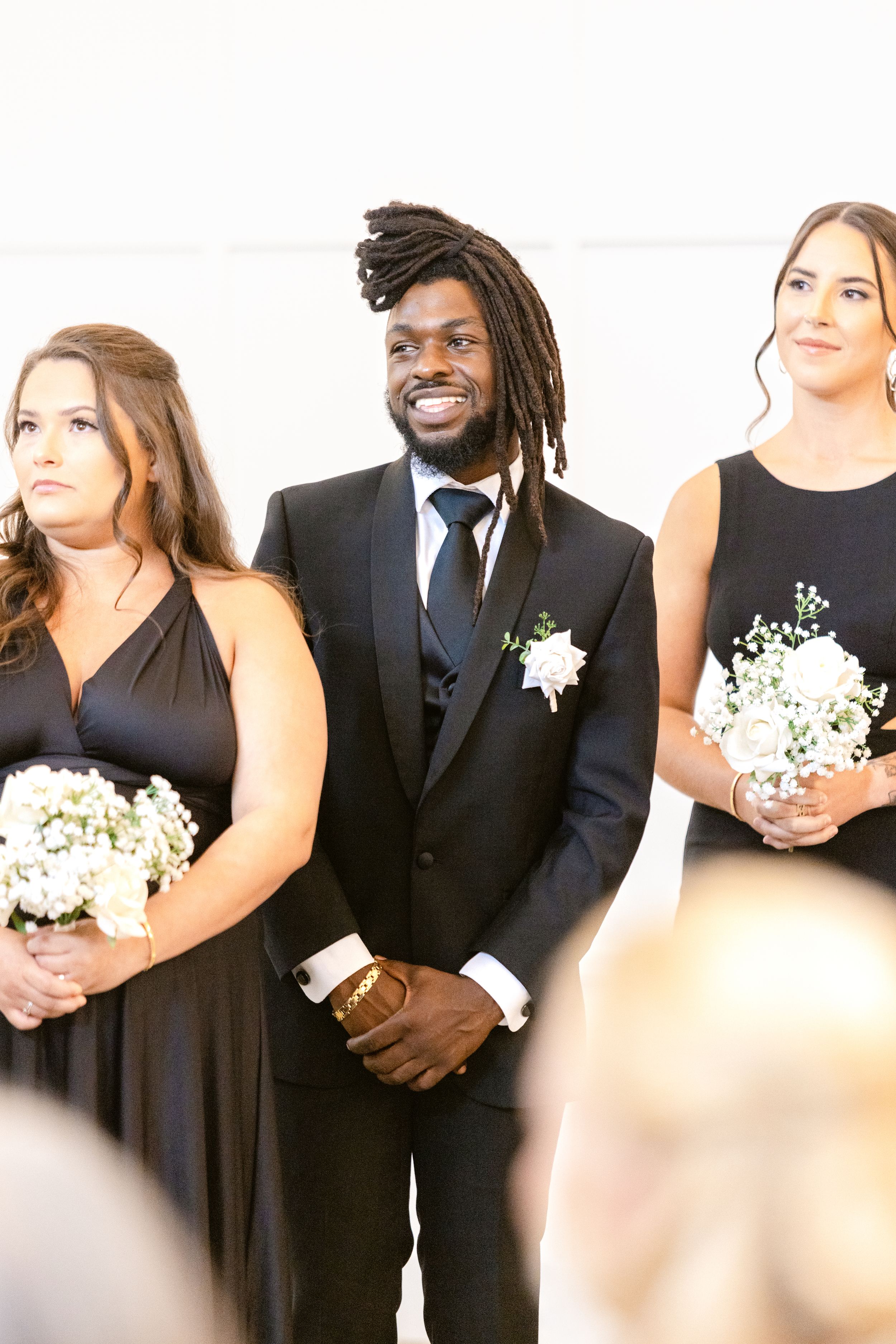 Wedding party members in black attire and white dresses pose together holding white floral bouquets.