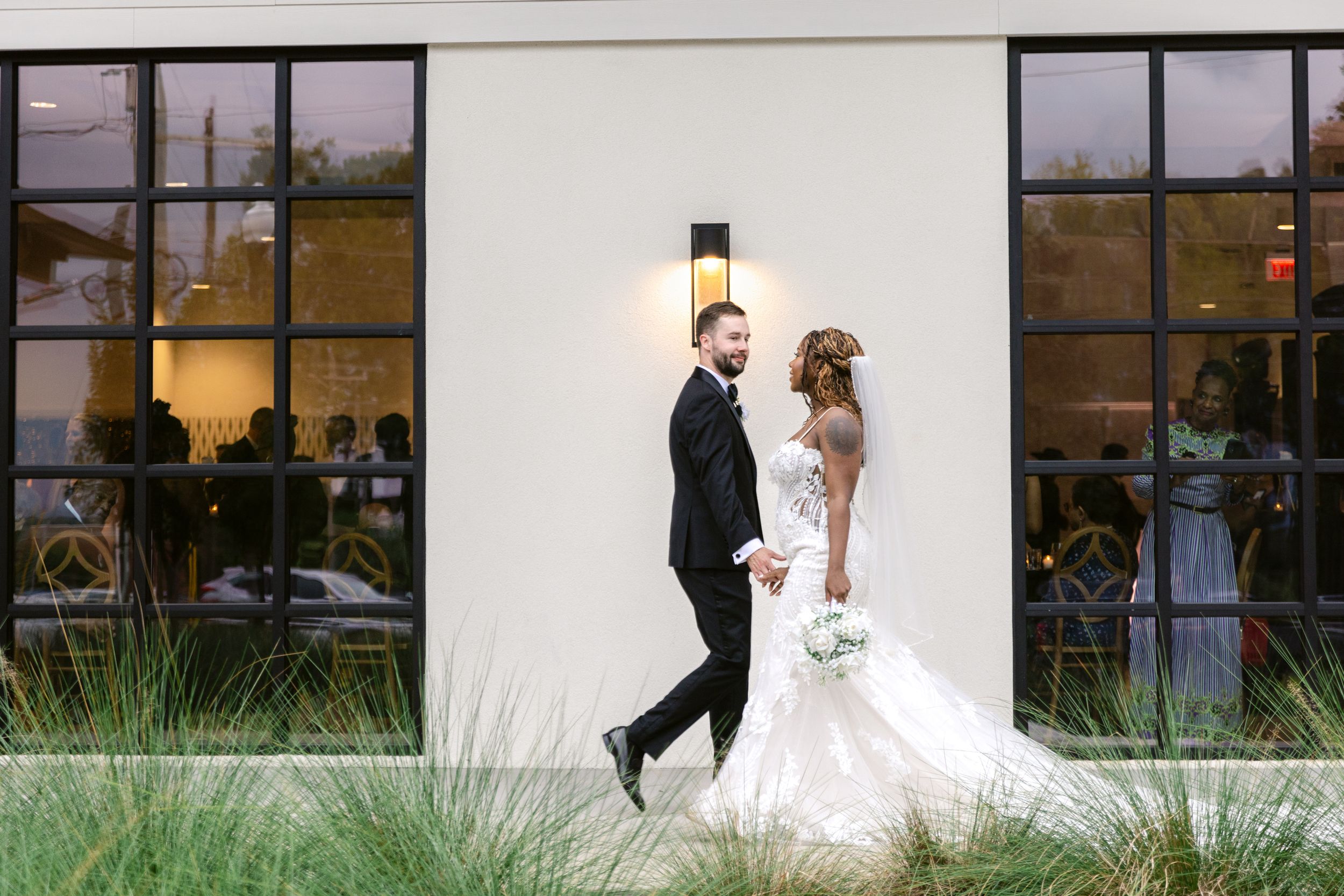 A wedding couple walks together along a modern building exterior with large windows at sunset.
