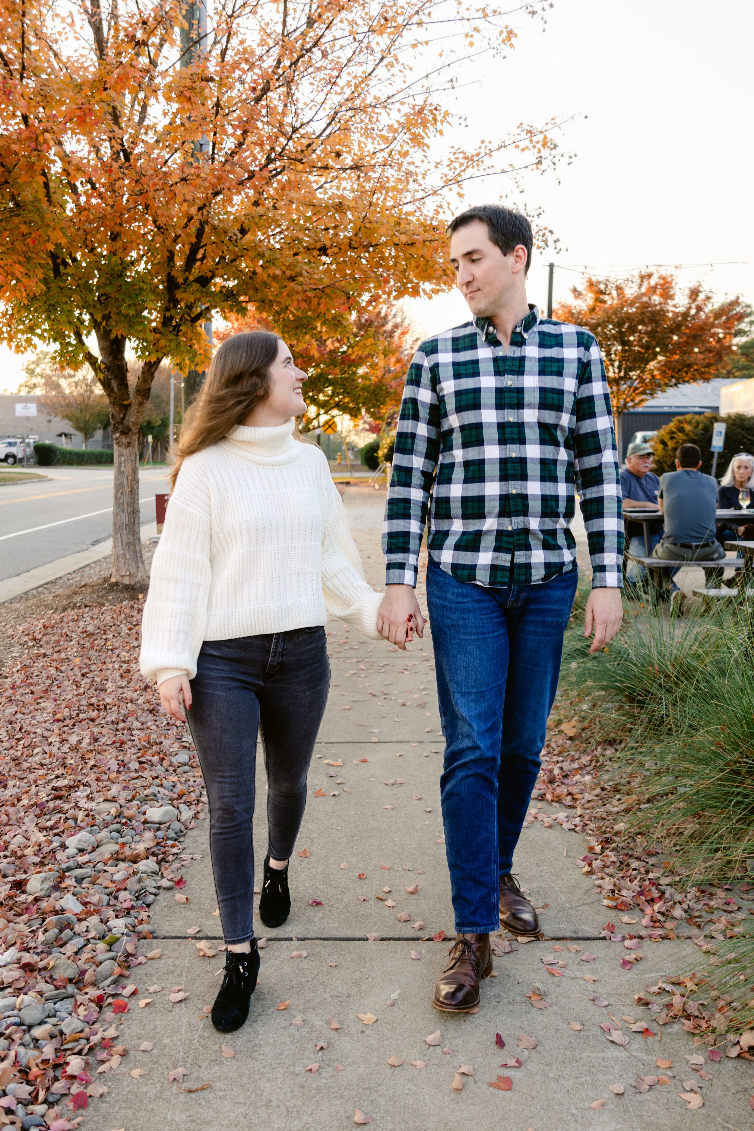 A couple takes a romantic autumn stroll on a sidewalk lined with orange trees and fallen leaves.