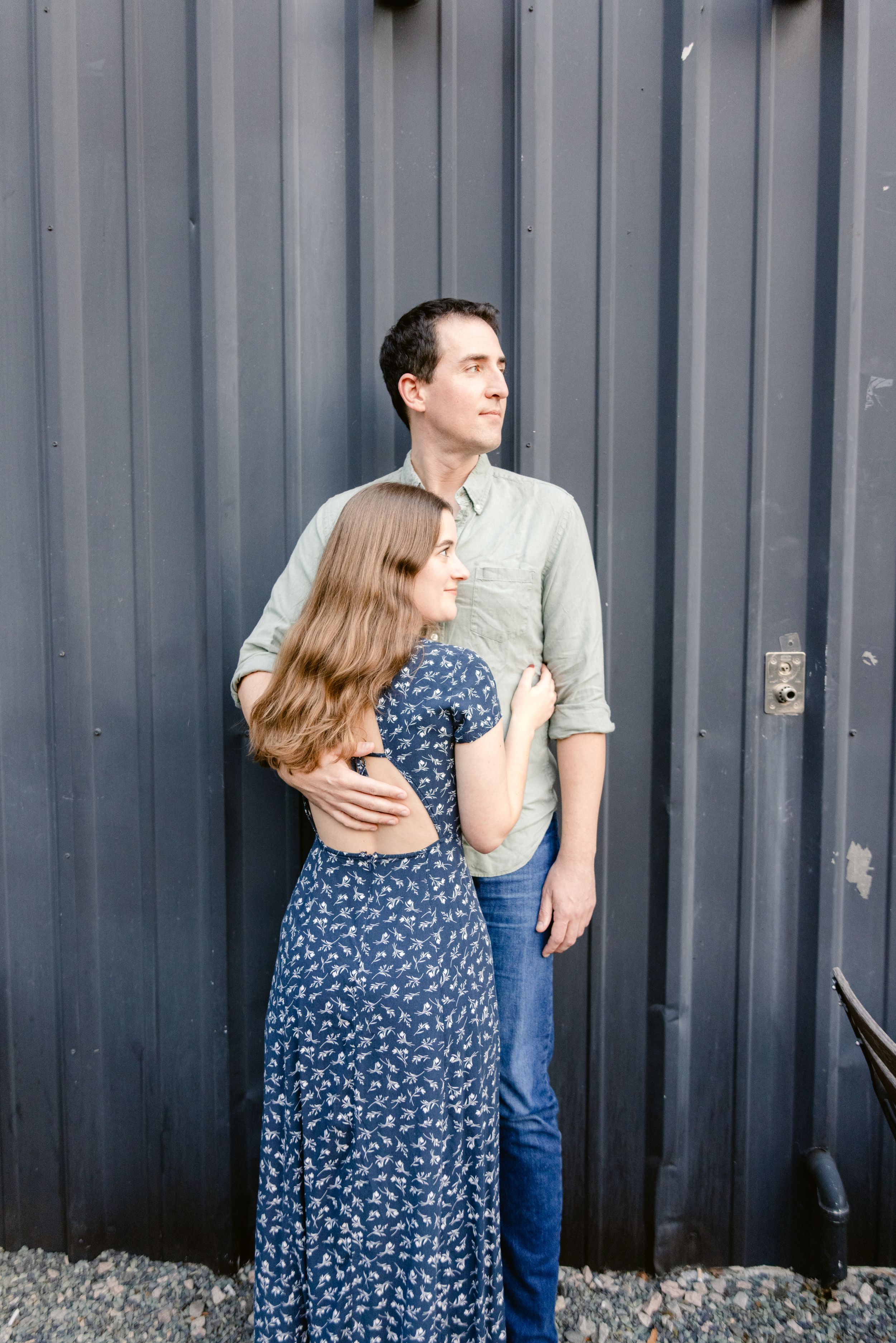 A couple embraces while standing against a grey corrugated metal wall.