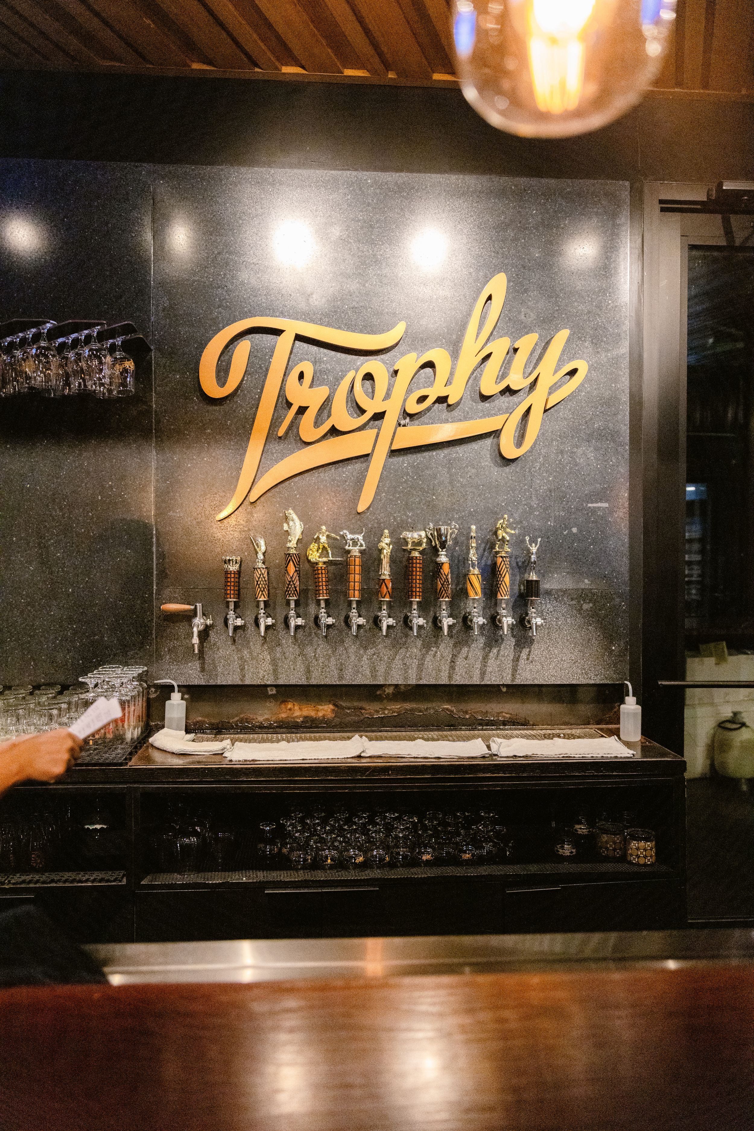 Trophy Brewing beer tap wall featuring illuminated gold lettering and multiple draft handles mounted on dark background.
