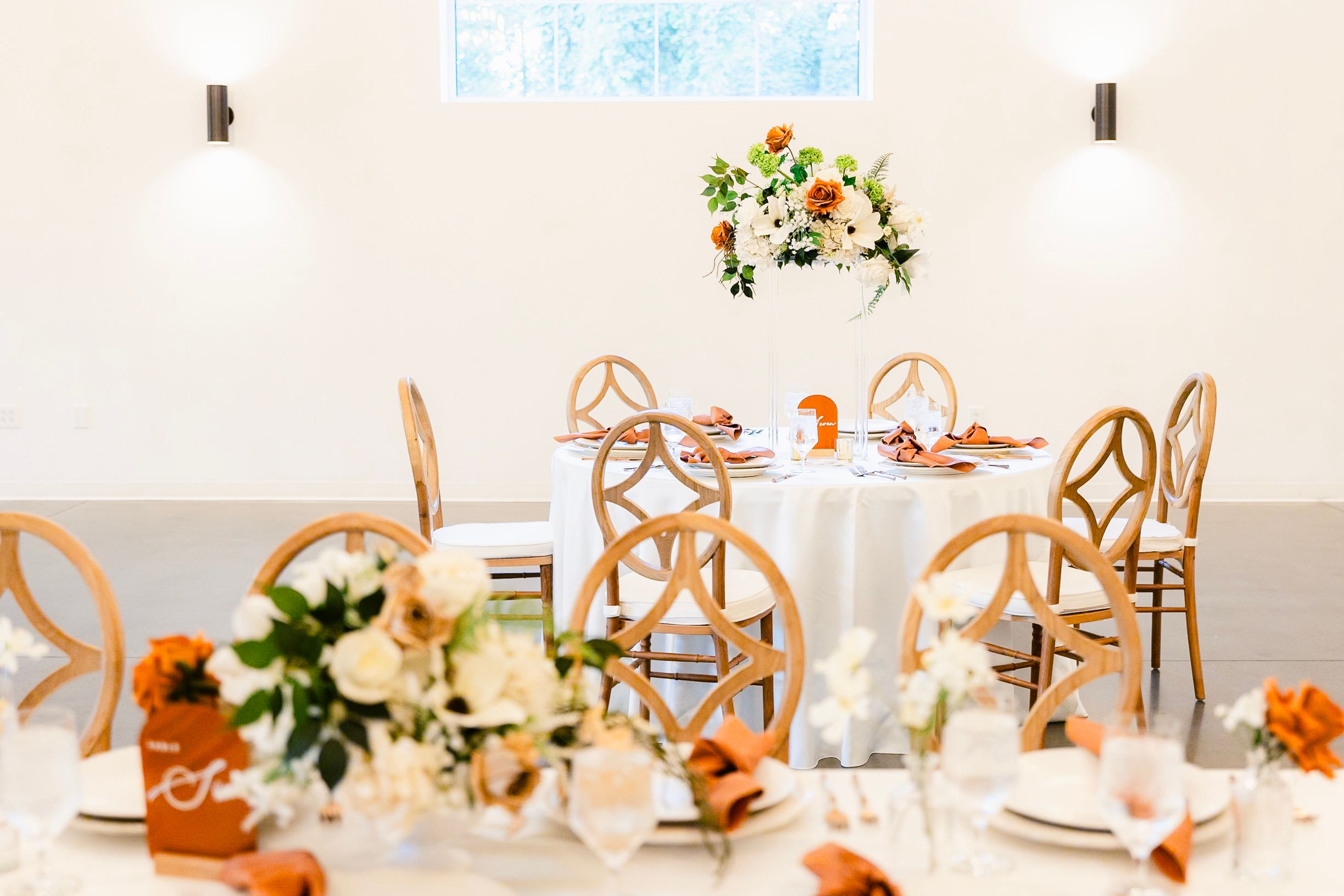 Elegant wedding reception table setting with wooden chairs and peachy floral centerpiece in a bright white room.