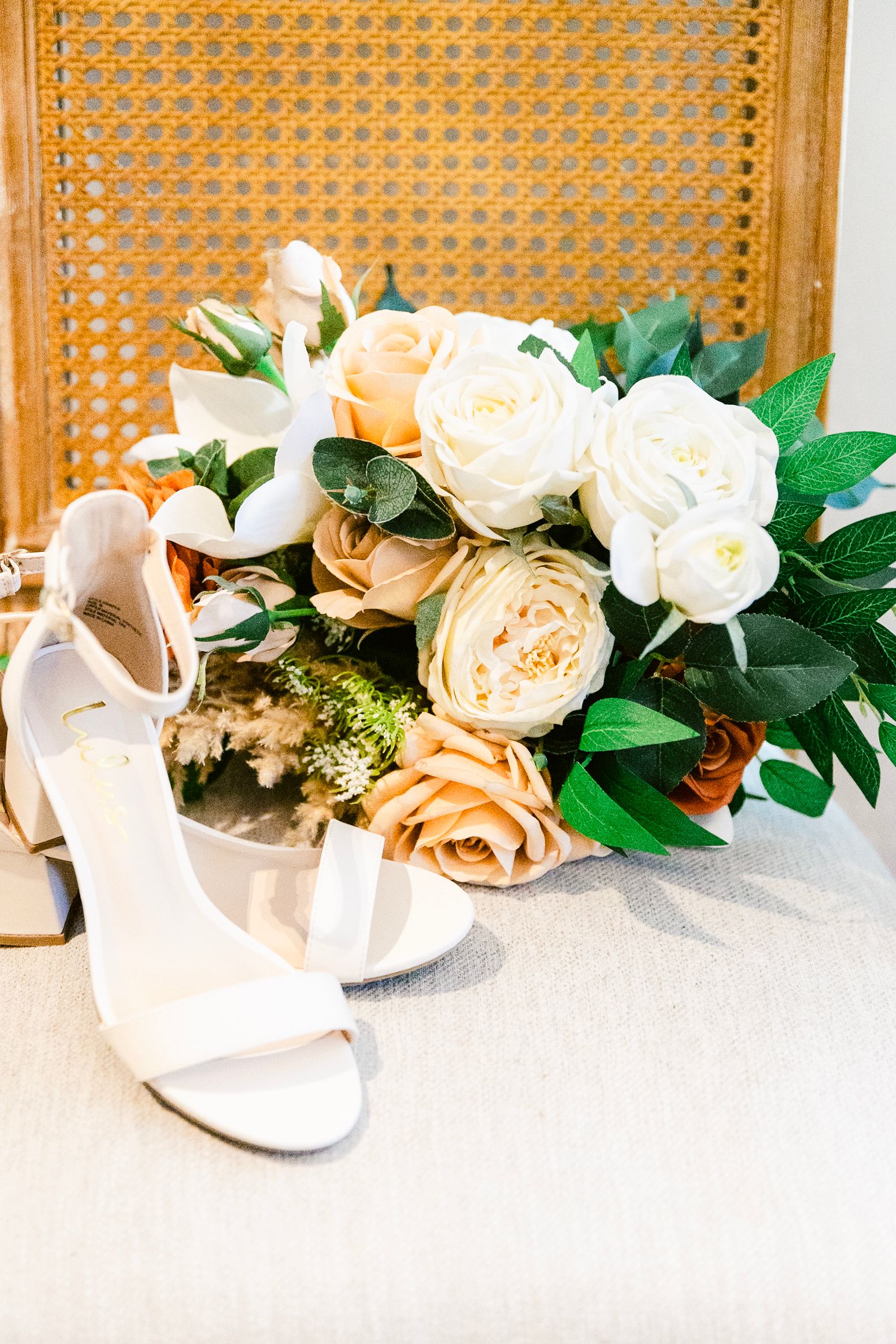 A beautiful bridal bouquet of peach and cream roses with greenery sits next to white wedding shoes.
