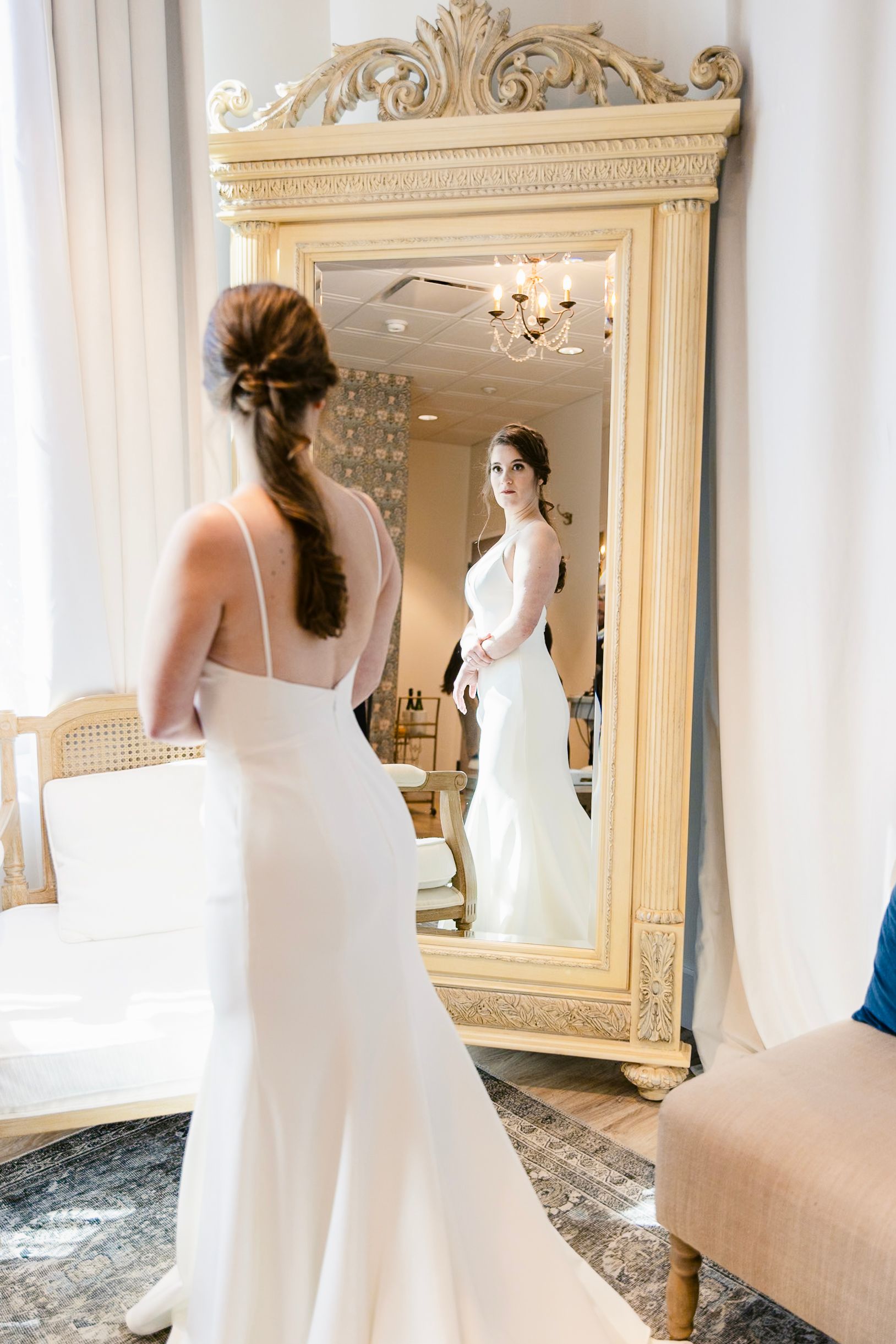 A bride in a white wedding gown looks at her reflection in an ornate cream-colored full length mirror.