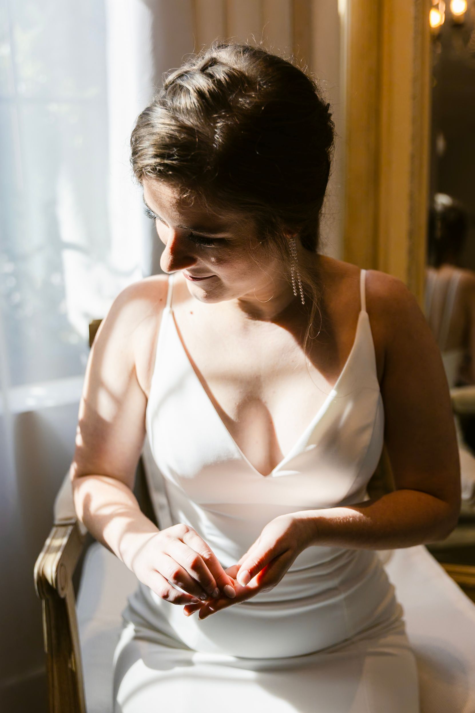 A bride in a simple white satin slip dress sits by a window in soft natural light.