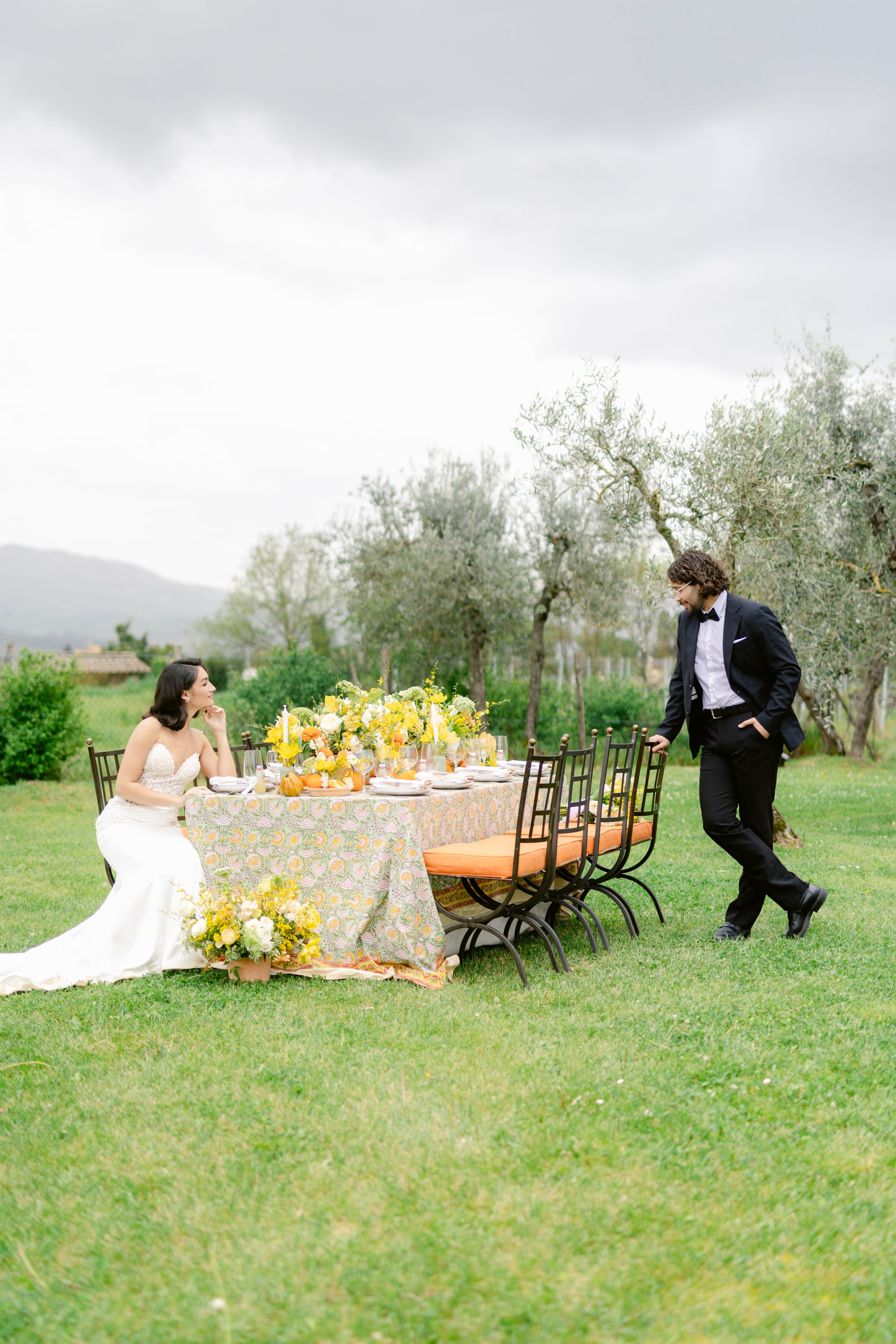 An elegant outdoor wedding table setting with yellow floral decor and iron chairs in a Tuscan olive grove.