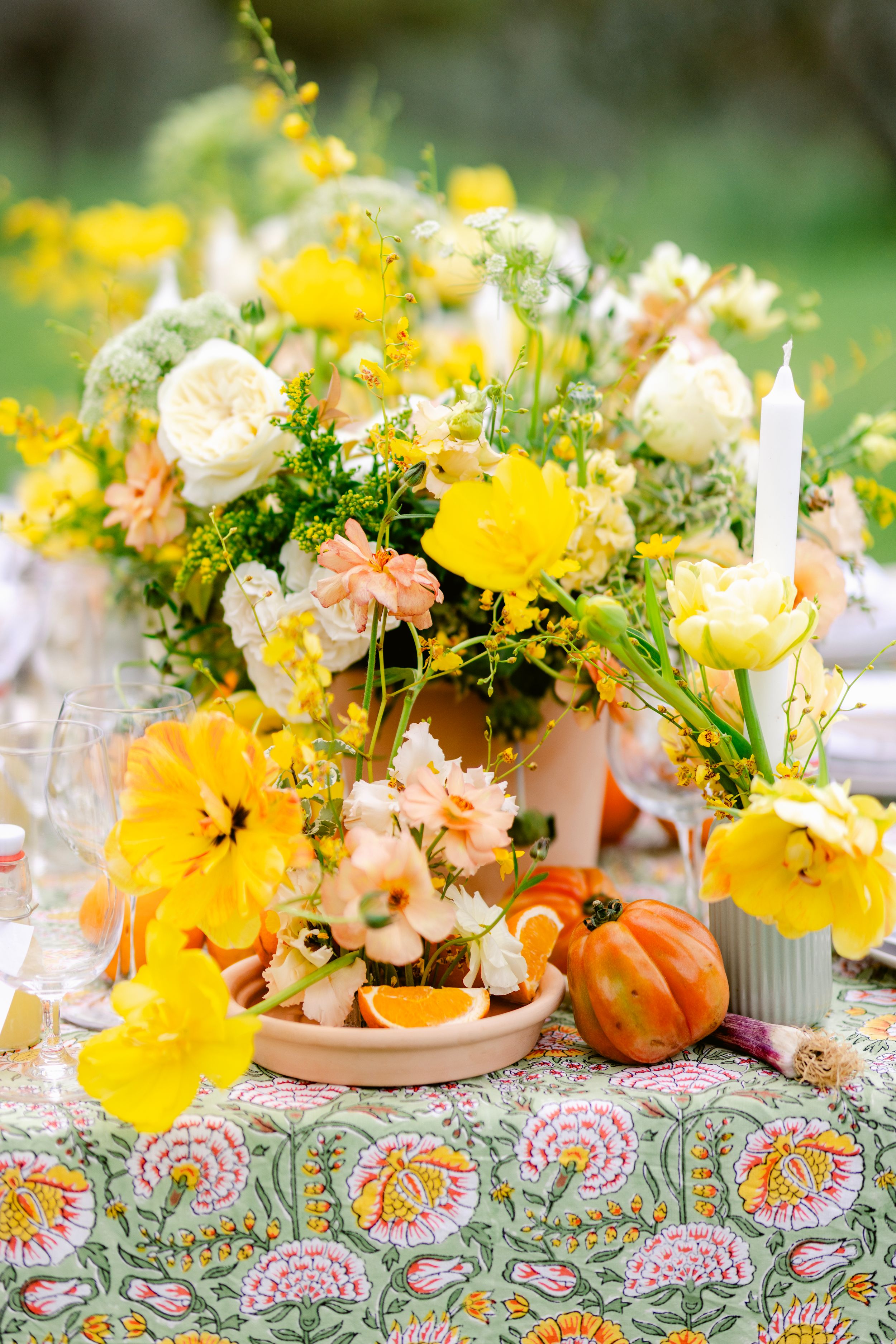 Fall table decor features bright yellow and peach floral centerpieces with small pumpkins on a colorful patterned tablecloth.