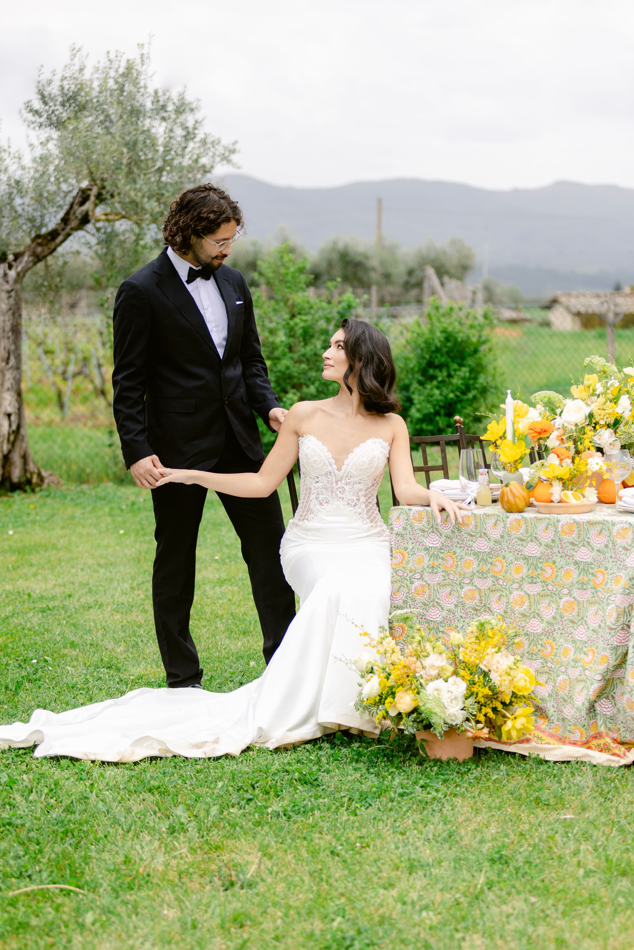 Couple in wedding attire having a romantic picnic outdoors with yellow floral decor and mountain views.
