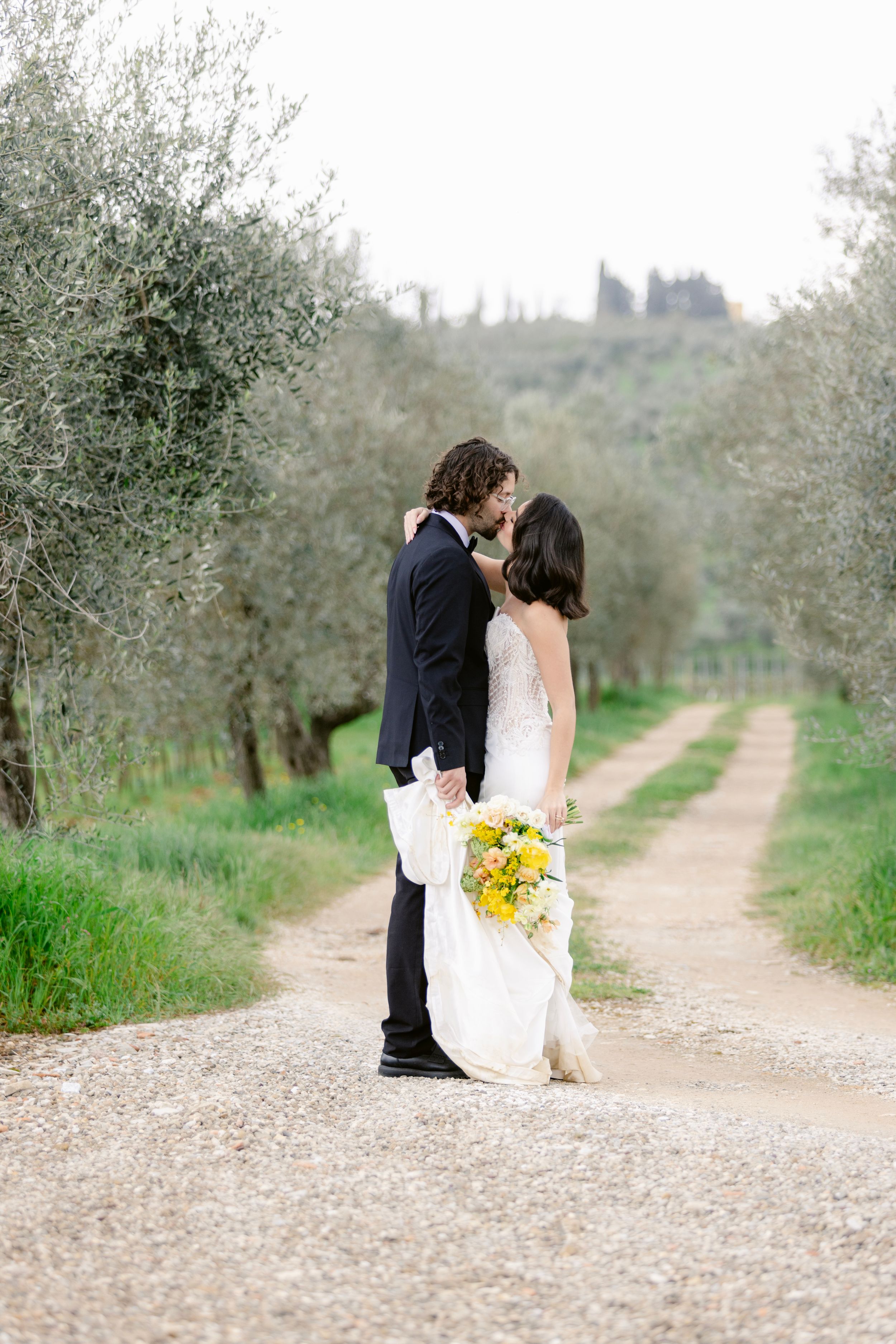 A romantic wedding moment captured on a rustic path lined with olive trees in the Italian countryside.