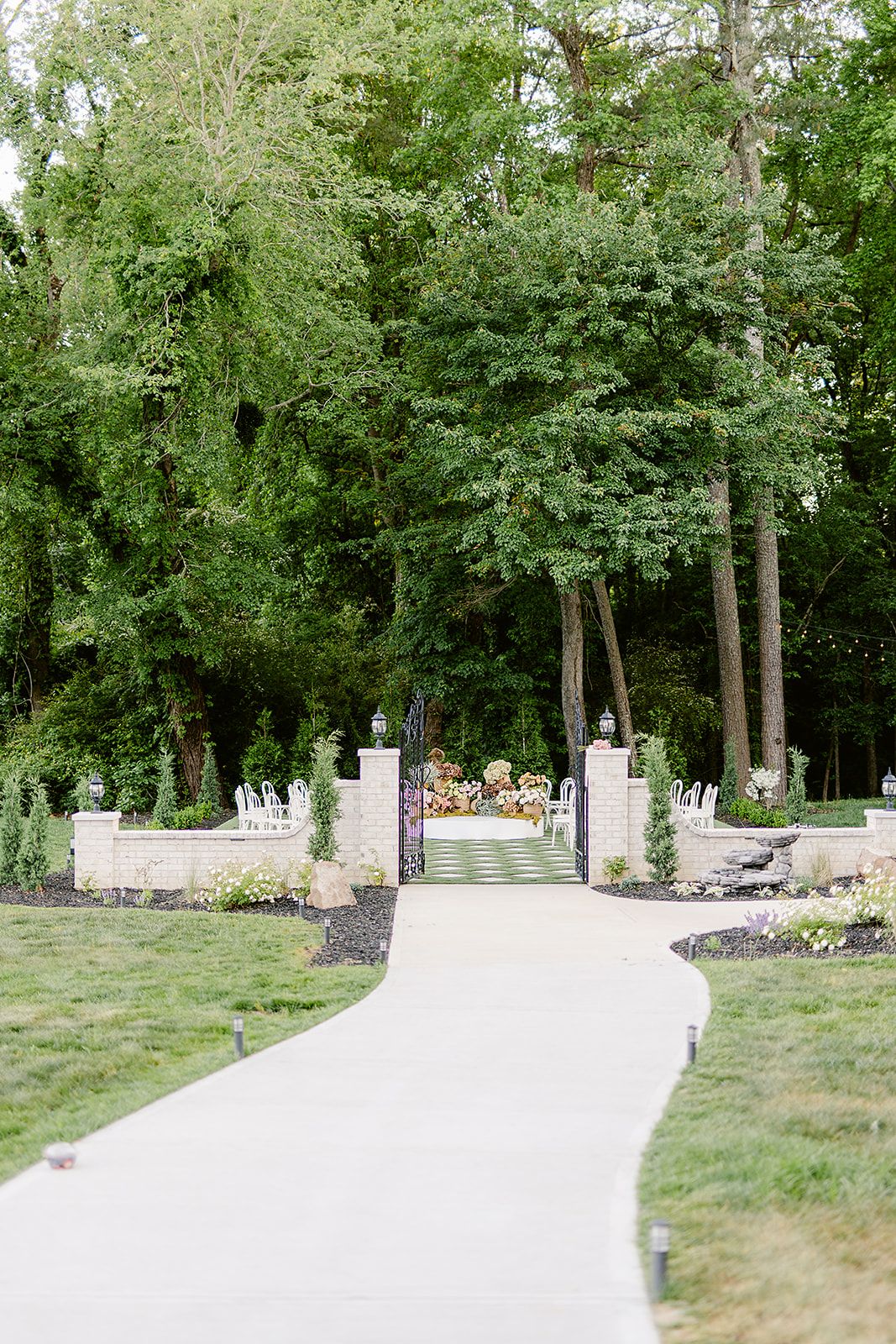 White stone pathway leading to elegant pillared entrance gates surrounded by lush green trees.