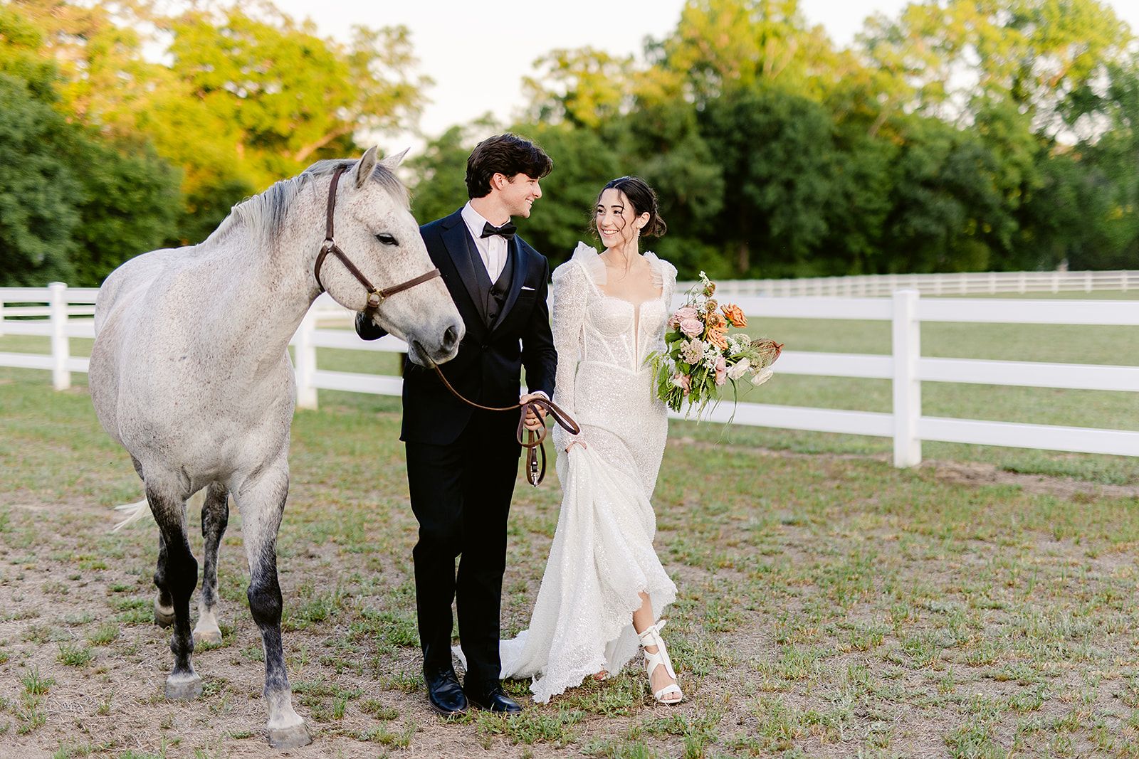 A couple in wedding attire walks with a white horse along a fence-lined path at an outdoor venue.