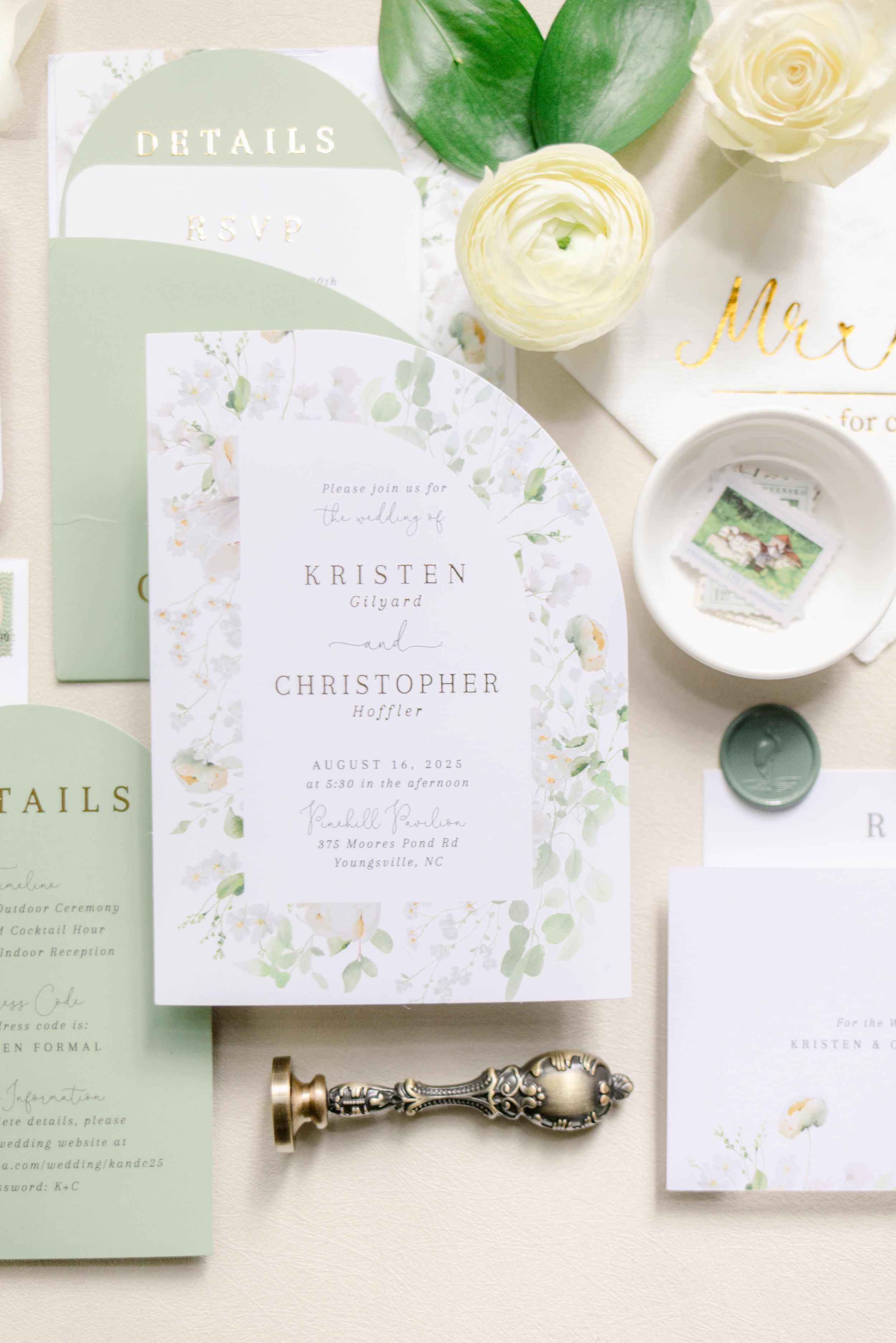 Elegant wedding invitation suite in white and sage green with floral details and gold accents laid out on cream surface.