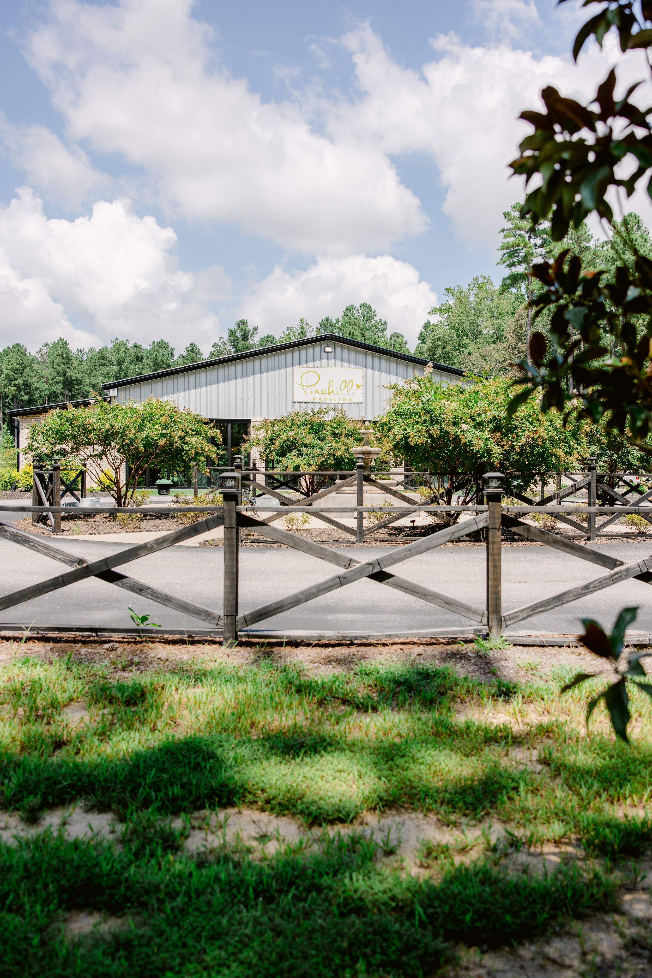 Wooden cross-beam fencing stretches along a grassy field with trees and a white building in the background.