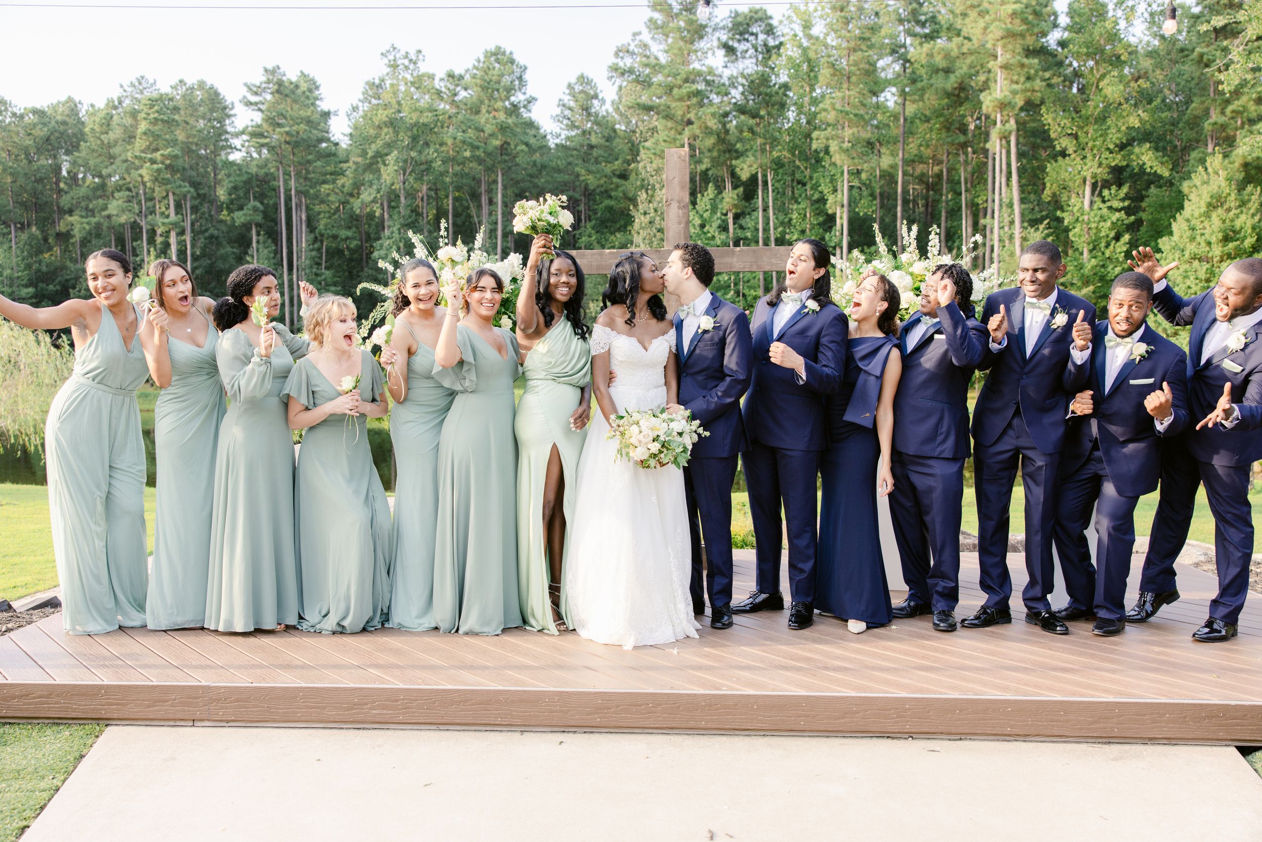 Wedding party members in navy suits and sage green dresses pose together outdoors for formal group photos.